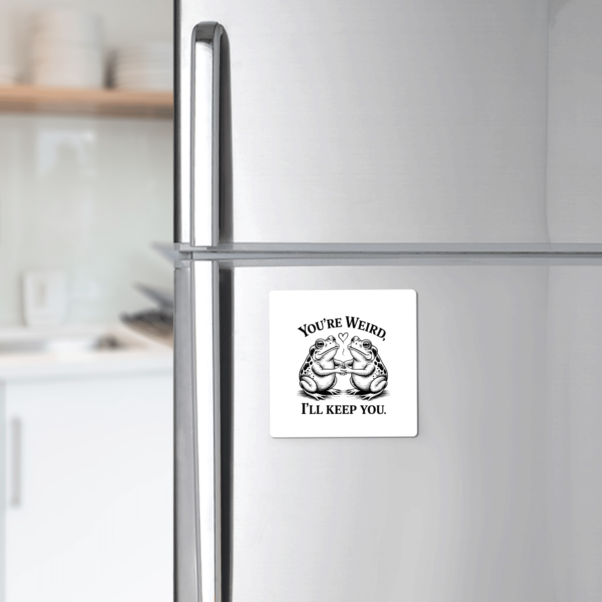 A refrigerator adorned with the Mirabilia Boutique "You're Weird, I'll Keep You" Frog Magnet—a quirky gift featuring a pair of hugging frogs. In the softly blurred background, shelves stacked with plates enhance the cozy cottagecore ambiance.