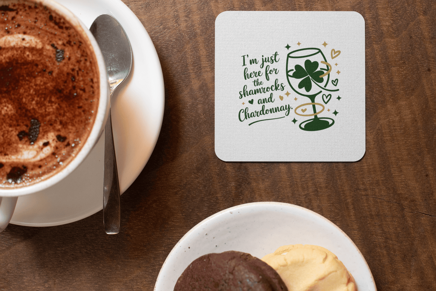 A cup of hot chocolate with a saucer, two cookies on a plate, and the Mirabilia Boutique Shamrocks and Chardonnay Coaster Set—featuring green shamrock and wine glass illustrations—a perfect gift for wine lovers.