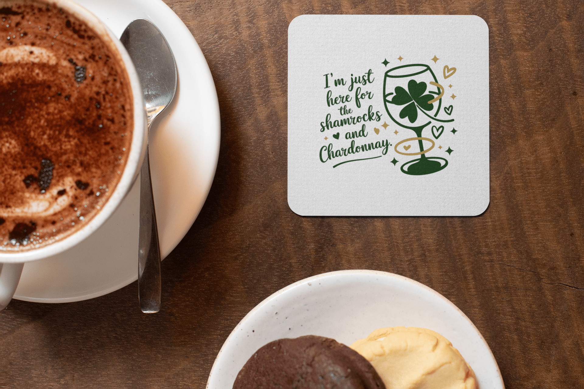 A cup of hot chocolate with a saucer, two cookies on a plate, and the Mirabilia Boutique Shamrocks and Chardonnay Coaster Set—featuring green shamrock and wine glass illustrations—a perfect gift for wine lovers.