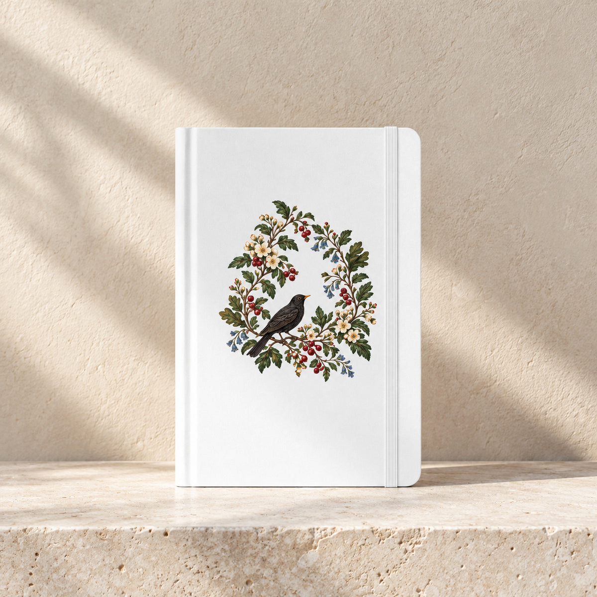 The Garden Ledger A5 Lined Journal by Mirabilia Boutique, featuring a white cover with a central illustration of a black bird on a branch, surrounded by a botanical wreath of flowers and berries.