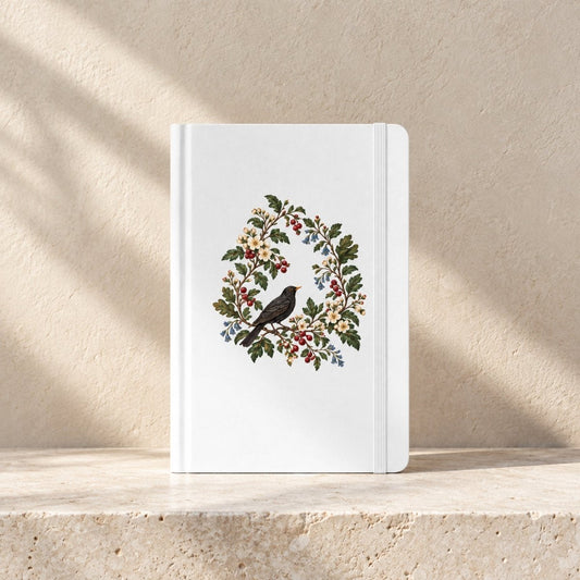 The Garden Ledger A5 Lined Journal by Mirabilia Boutique, featuring a white cover with a central illustration of a black bird on a branch, surrounded by a botanical wreath of flowers and berries.