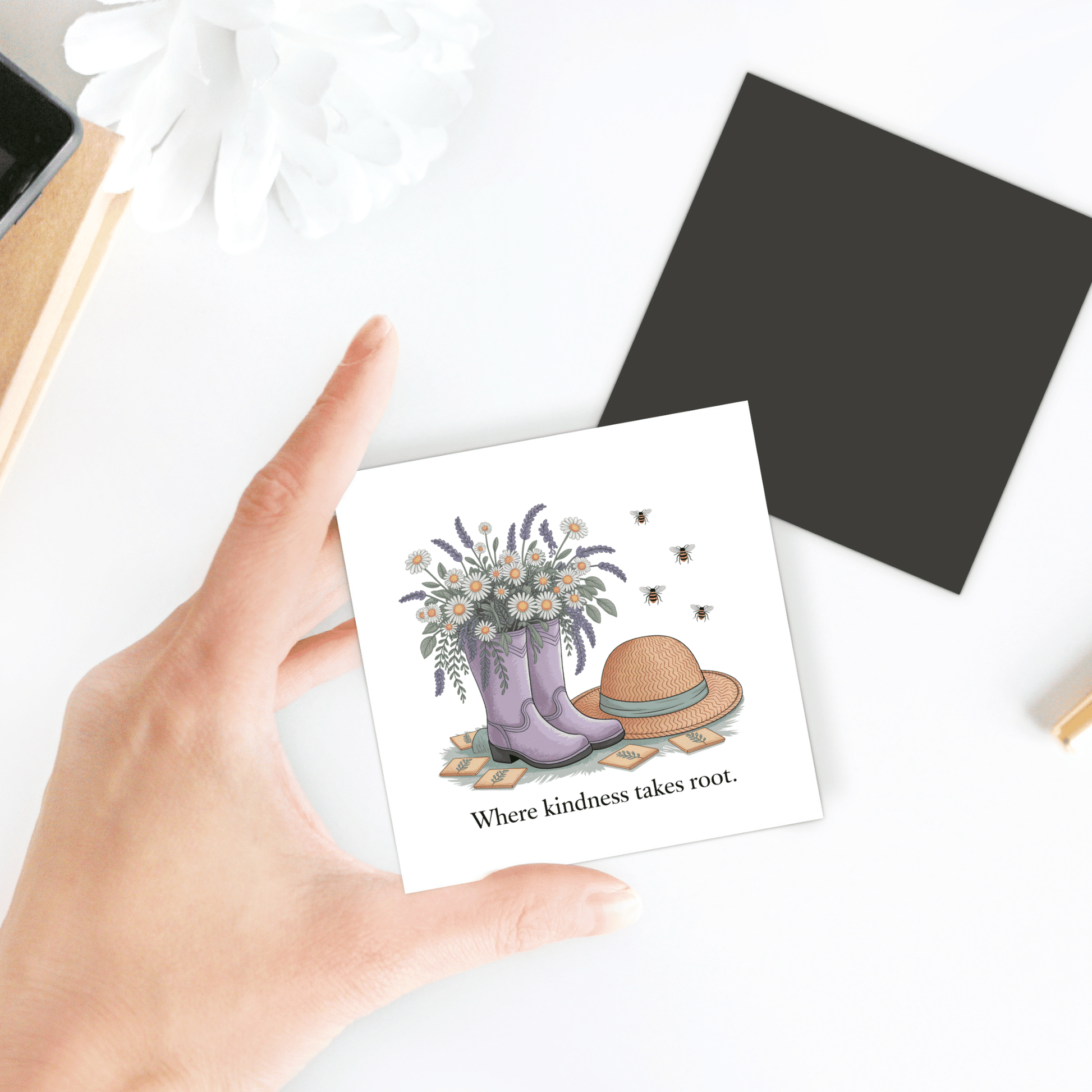 A hand holds the Mirabilia Boutique Kindness Garden Magnet – Floral Boots & Bees Quote, featuring wildflowers in purple boots, a straw hat, bees, and the phrase "Where kindness takes root." A charming kindness gift.