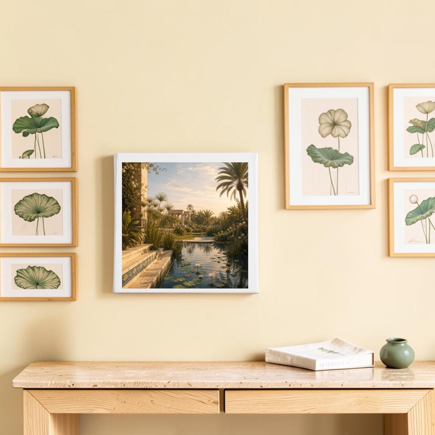 A minimalist wooden console table sits beneath six botanical prints and a central framed photo of a tranquil garden with palms and water, all set against a light beige wall. To one side, the “Garden of Still Waters | Egyptian Canvas Wall Art” by Mirabilia Boutique—a refined lotus blossom painting inspired by ancient Egypt—adds an elegant historical touch. A book and a small dish rest on the table.