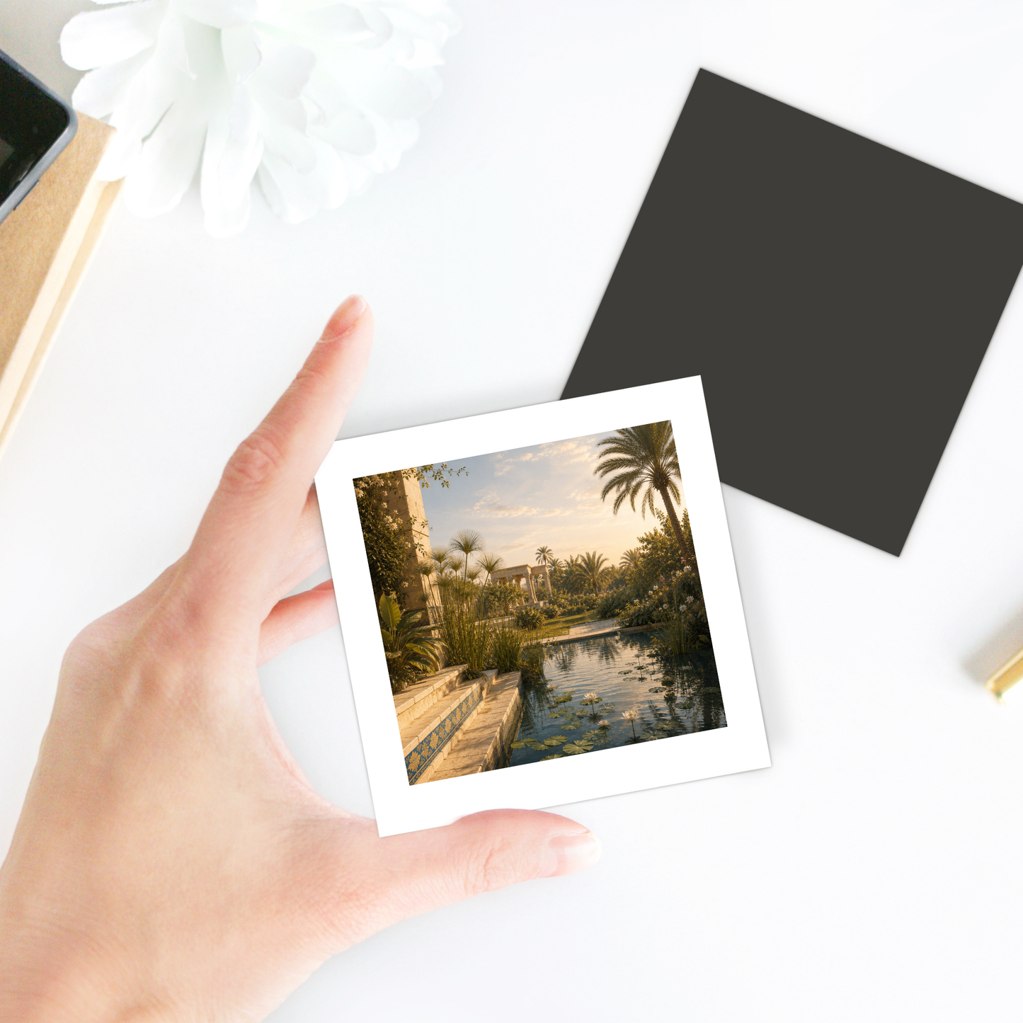A hand holds a photo of a lush garden with palm trees, a pond, and classical stone steps, evoking the charm of ancient Egypt decor. Other items—including a flower and a black square card featuring the Garden of Still Waters Magnet by Mirabilia Boutique—are arranged on a white table in the background.
