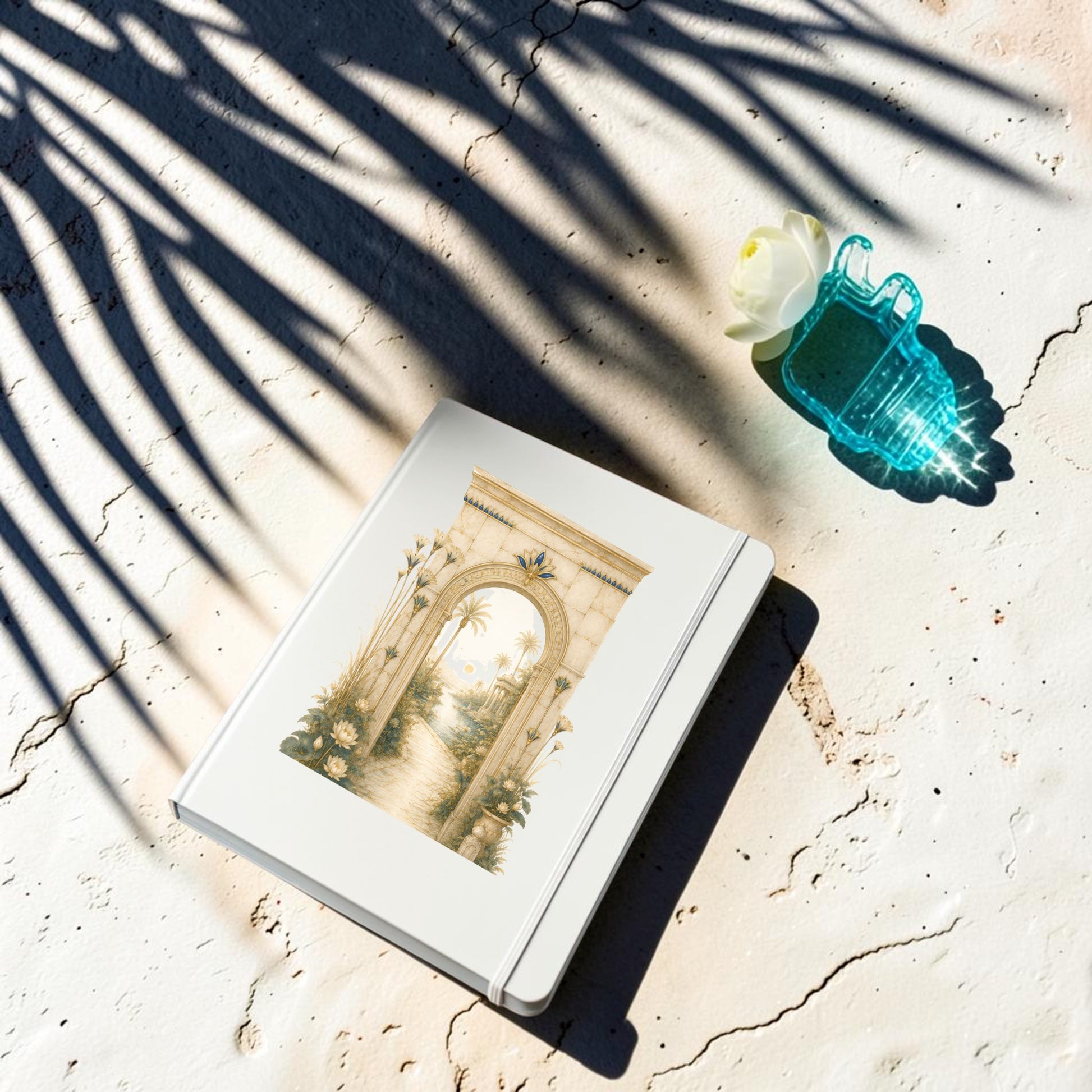 The Garden Path Journal from the Lapis & Lotus Collection by Mirabilia Boutique, inspired by ancient Egypt and adorned with an archway illustration, a blue transparent lock, and a white flower, rests on a sunlit, textured surface with palm leaf shadows.