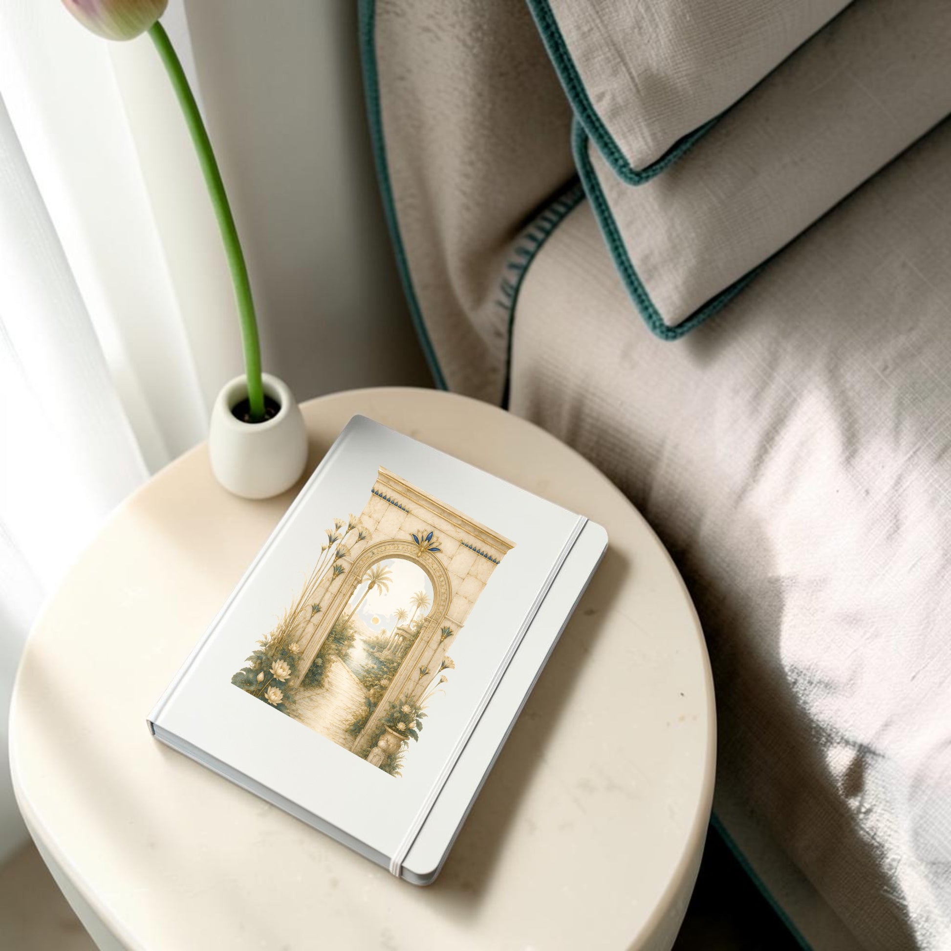 The Garden Path Journal | Ancient Egyptian Inspired Notebook from Mirabilia Boutique, adorned with an archway and garden illustration from the Lapis & Lotus Collection, rests on a round bedside table beside a bed and a white vase holding a single tulip.