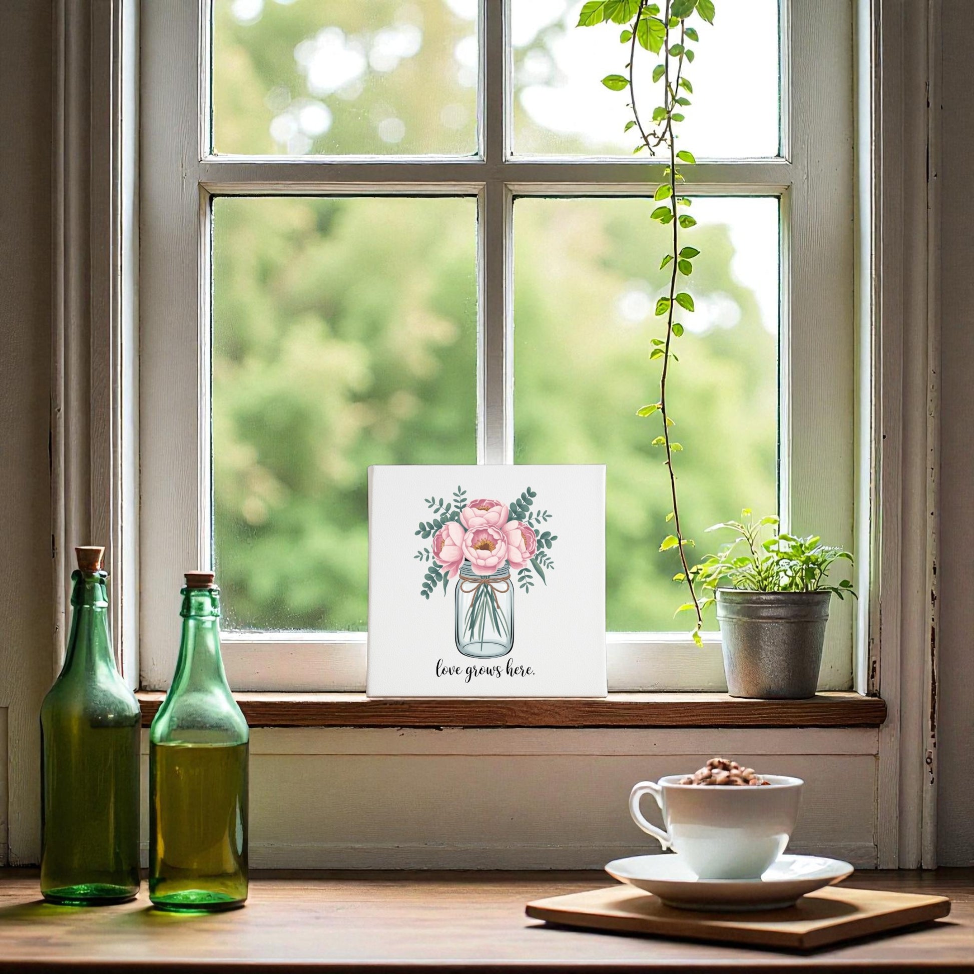 A cozy windowsill with green glass bottles, a potted plant, and cocoa, featuring Mirabilia Boutique's Love Grows Here Floral Canvas Wall Art, surrounded by trees and gentle blush peonies wall art visible through the window.