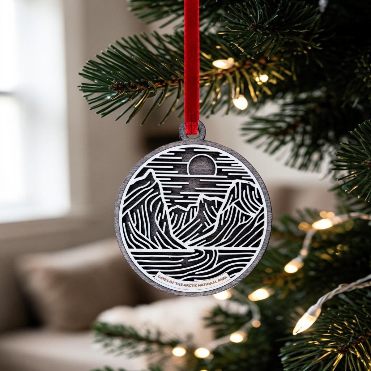 The Gates of the Arctic Line Art Christmas Ornament by Mirabilia Boutique, featuring a metal mountain and sun design, hangs from a red ribbon on a Christmas tree branch adorned with white lights; in the background, a blurred couch and window are visible.