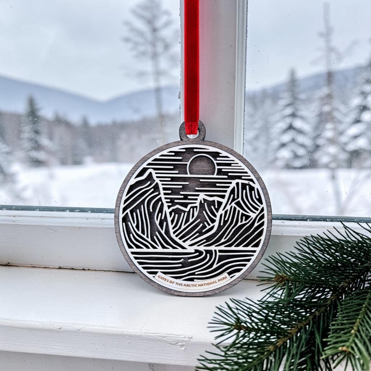 The Gates of the Arctic Line Art Christmas Ornament by Mirabilia Boutique, featuring a black-and-white mountain scene, hangs by a red ribbon in front of a snowy window; pine branches peek from the corner, capturing the rugged beauty of Alaska’s Gates of the Arctic National Park.