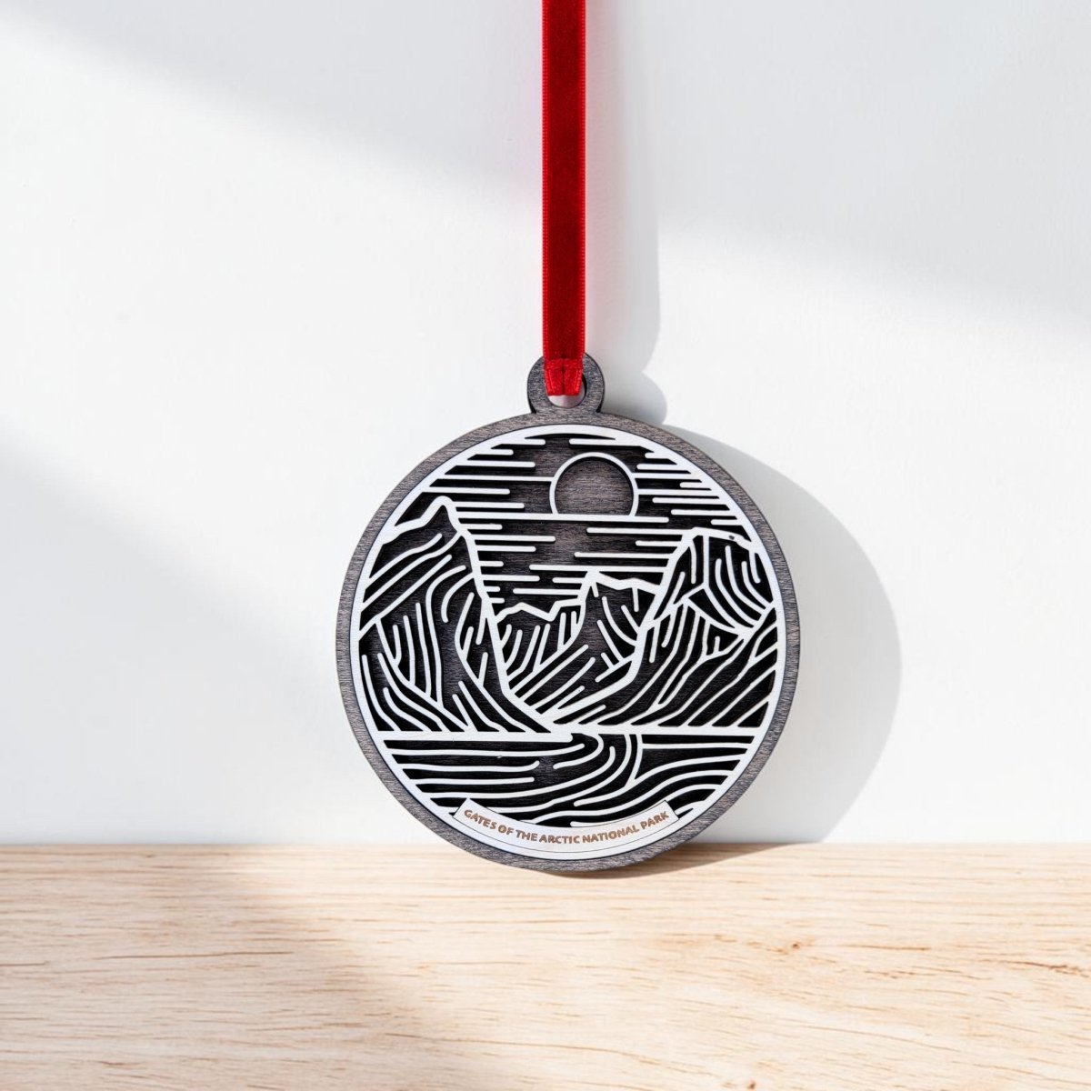 The Gates of the Arctic Line Art Christmas Ornament by Mirabilia Boutique, featuring a black and white mountain landscape design, hangs from a red ribbon against a white wall and light wooden surface—capturing the beauty of Alaska’s Gates of the Arctic National Park.