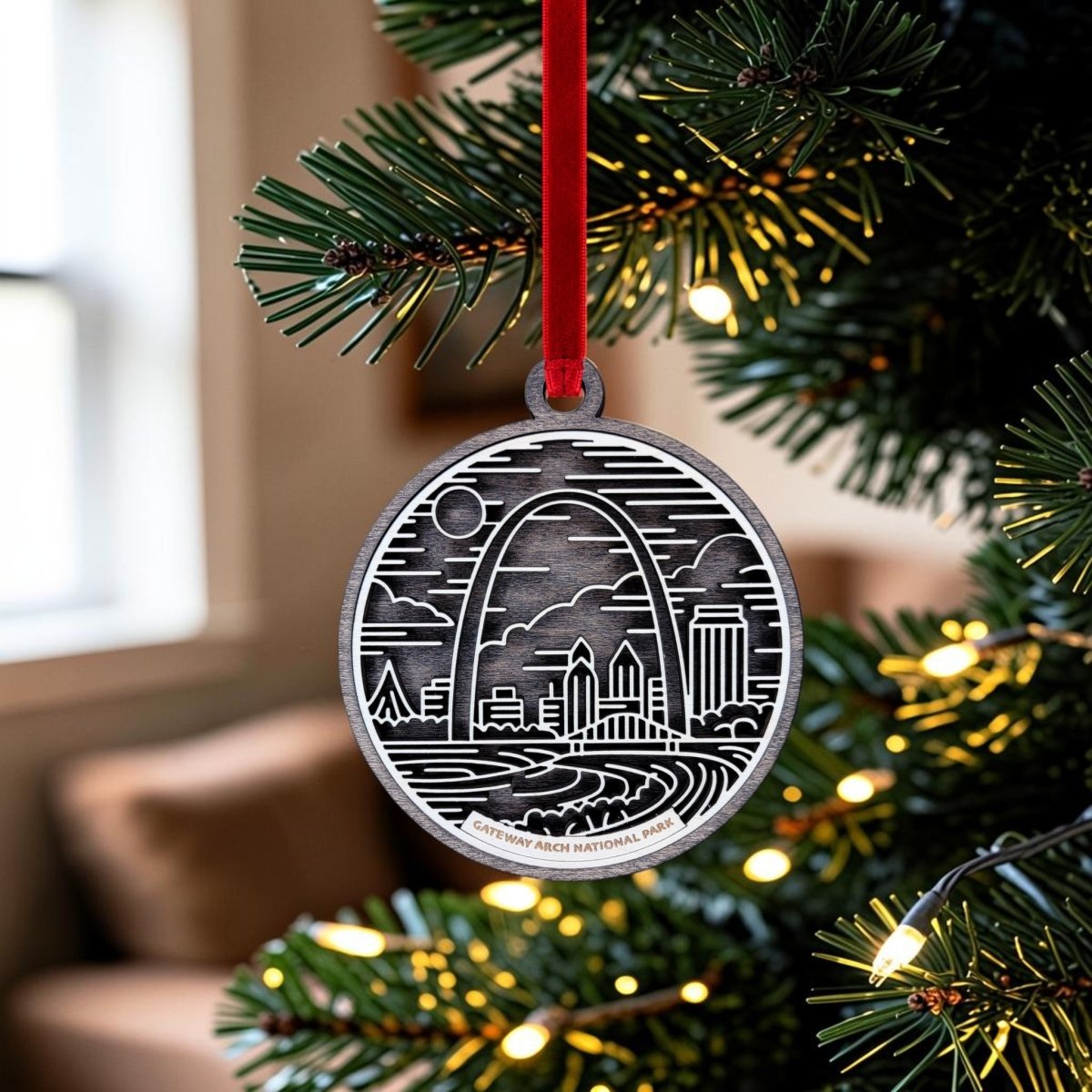 The Gateway Arch National Park Line Art Ornament by Mirabilia Boutique is a round silver ornament engraved with a detailed cityscape featuring the iconic St. Louis Gateway Arch, displayed here hanging from a red ribbon on a Christmas tree adorned with twinkling lights—a perfect St. Louis souvenir for the holiday season.