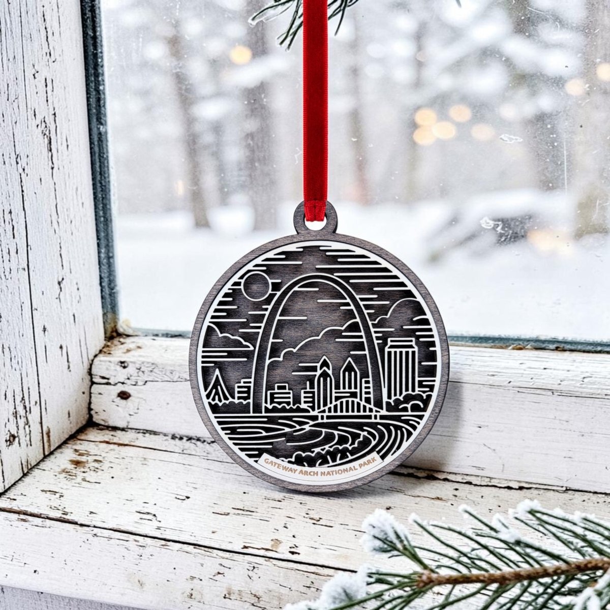 The Mirabilia Boutique Gateway Arch National Park Line Art Ornament features an engraved scene of the iconic Arch and city buildings, hanging by a red ribbon on a snowy window framed with evergreen branches and a wintry landscape outside—a perfect St. Louis souvenir that captures the spirit of Gateway Arch National Park.