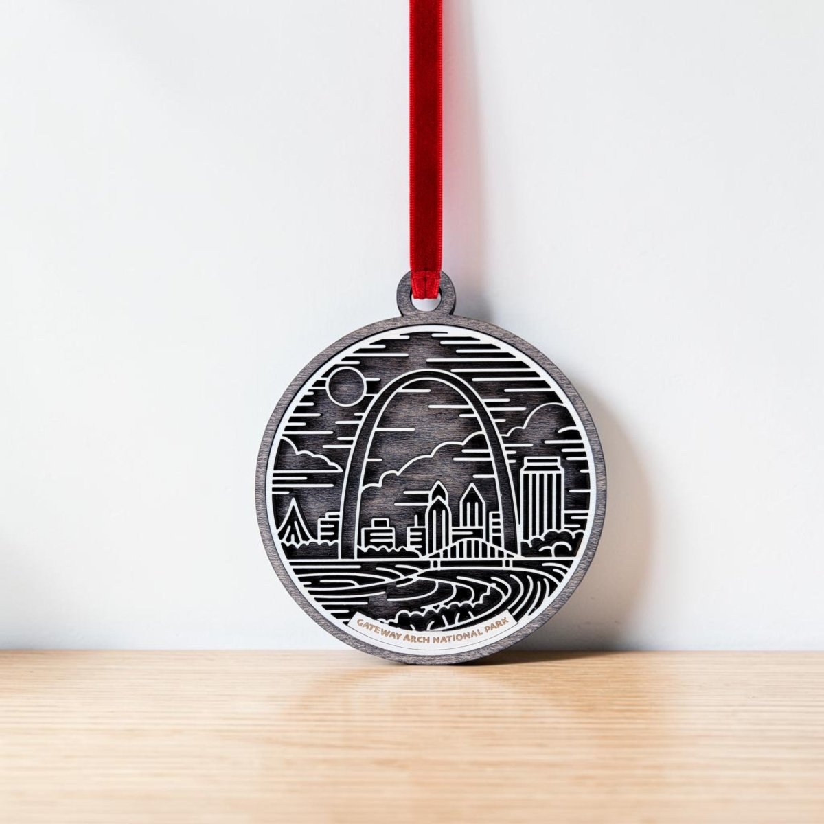 Bring home the perfect Gateway Arch ornament for any collection with the Gateway Arch National Park Line Art Ornament by Mirabilia Boutique. This round ornament features a line drawing of the iconic Arch, city buildings, clouds, and waves. Hung from a red ribbon against a white wall and wooden surface, it’s a charming St. Louis souvenir you’ll love to display.