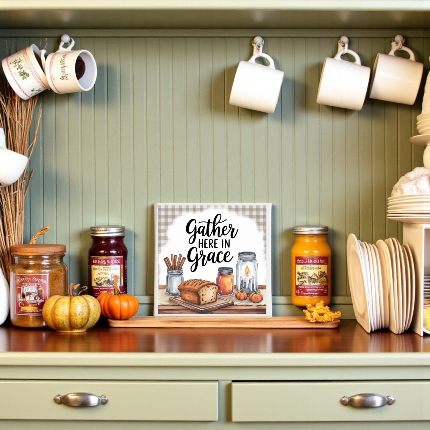 A kitchen shelf with mugs, jars of candles, plates, pumpkins, and the "Gather Here in Grace Canvas | 8x8 Farmhouse Kitchen Sign" by Mirabilia Boutique featuring bread and coffee illustrations on a green beadboard background.