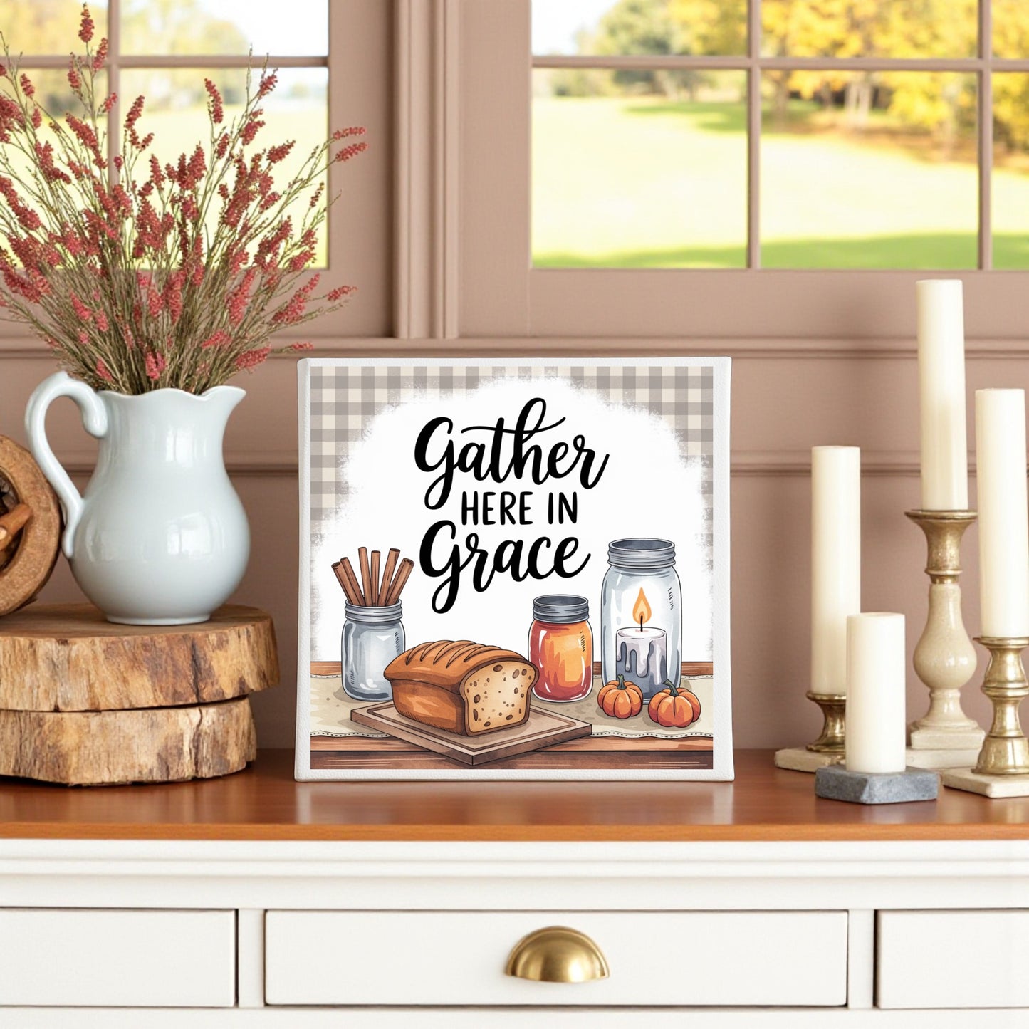 The Gather Here in Grace Canvas (8x8) by Mirabilia Boutique sits on a white dresser, surrounded by bread, candles, cinnamon sticks, jars, pumpkins, dried flowers in a vase, and brightened by a nearby window.