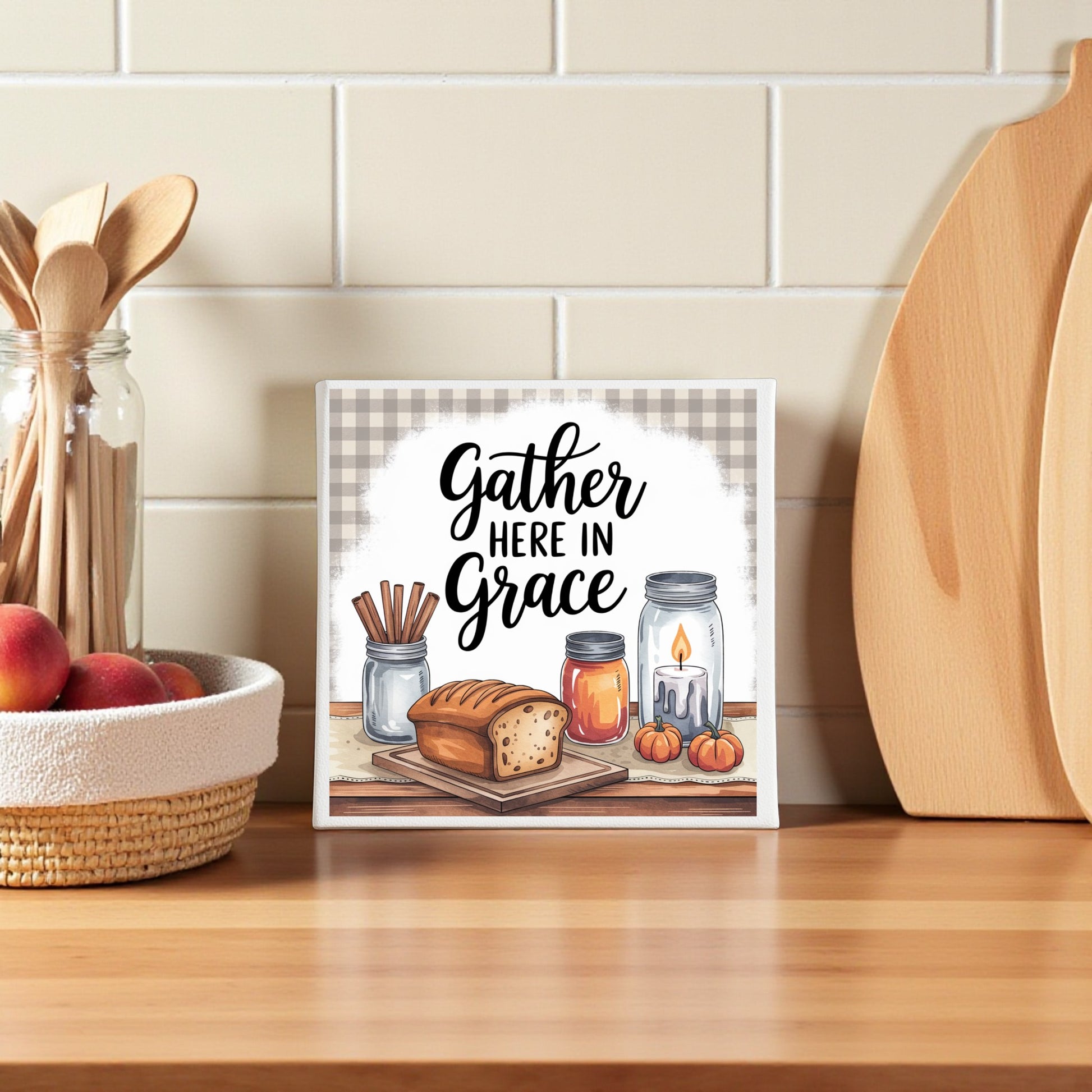 A Mirabilia Boutique "Gather Here in Grace" 8x8 canvas sign sits on the counter, surrounded by cinnamon jars, rustic bread, pumpkins, a candle, wooden utensils, and apples—ideal farmhouse kitchen decor.