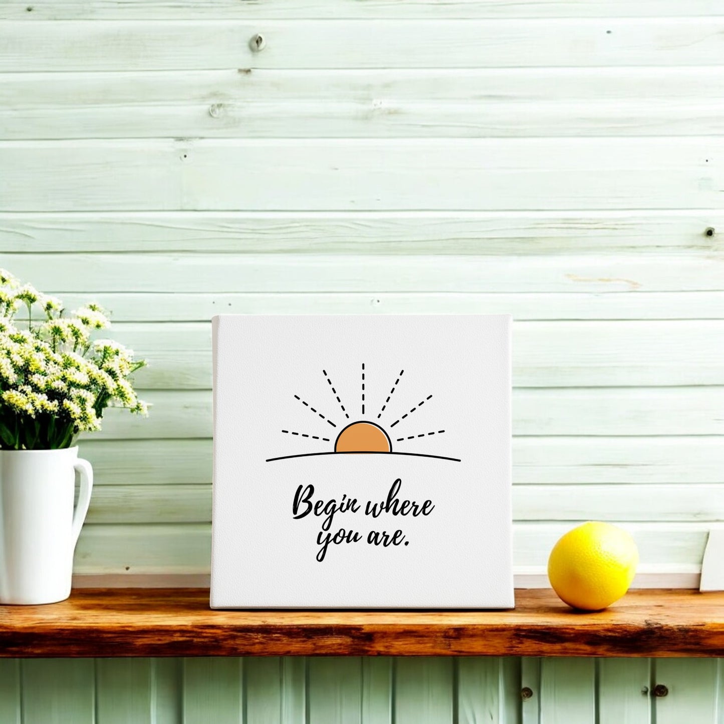 The Mirabilia Boutique "Begin Where You Are Canvas – Sunrise Wall Affirmation" sits on a wooden shelf with a vase of white flowers and a lemon, set against a light wood wall for a minimalist touch.