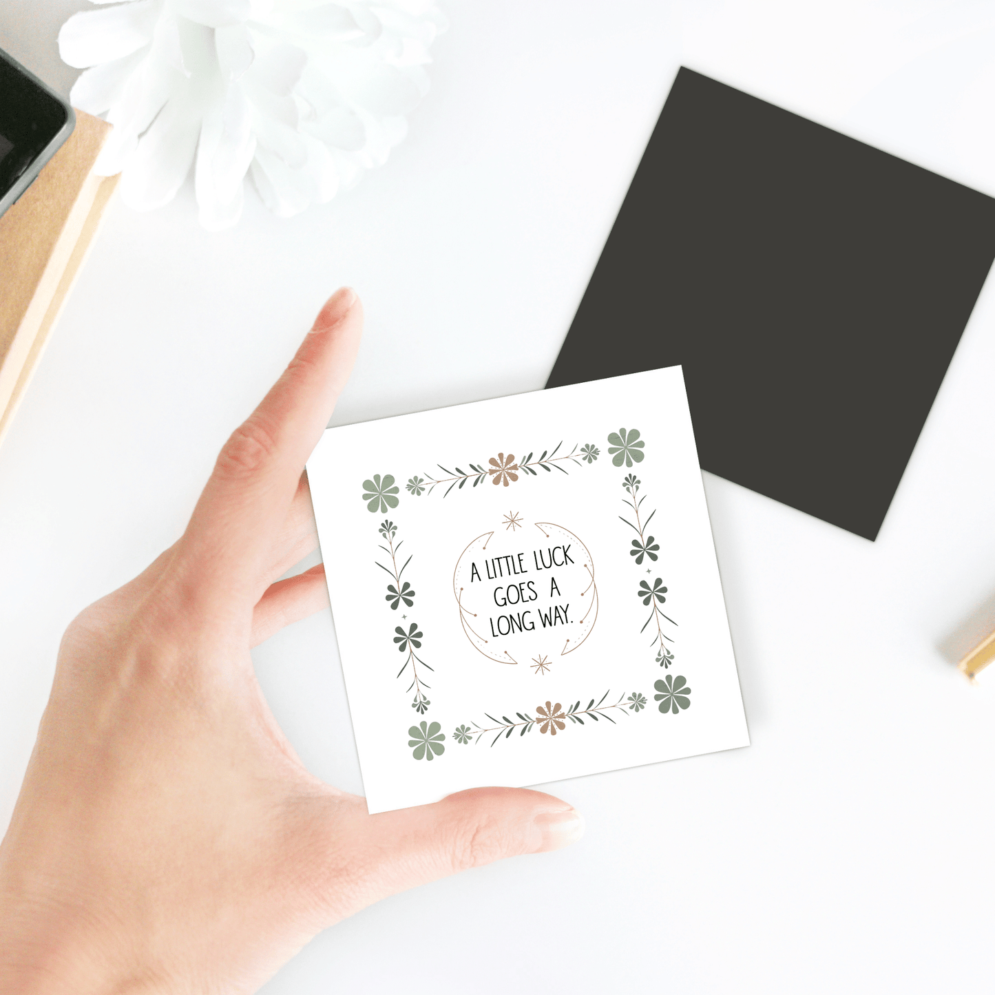 A hand holds Mirabilia Boutique’s “A Little Luck Goes a Long Way” magnet with green floral designs. In the background are a black card, notebook, and white flower on a white surface.