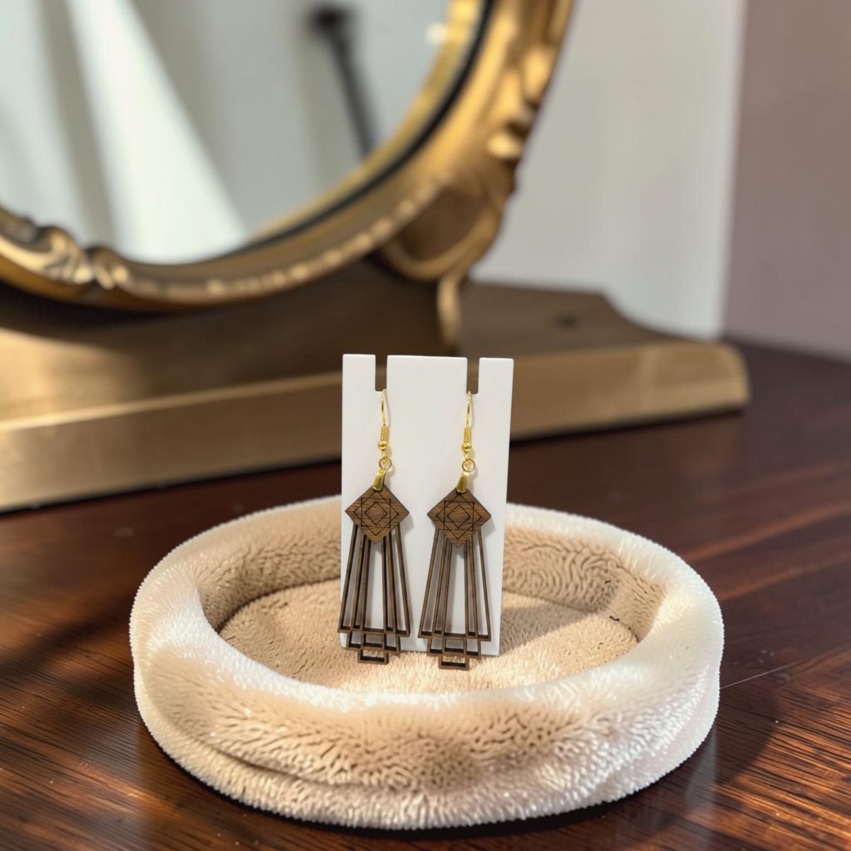 Art Deco Skyscraper Wood Statement Earrings by Mirabilia Boutique in rich brown tones with gold hooks are displayed on a white card, resting in a beige textured jewelry dish atop a wooden table, with a gold-framed mirror in the background.