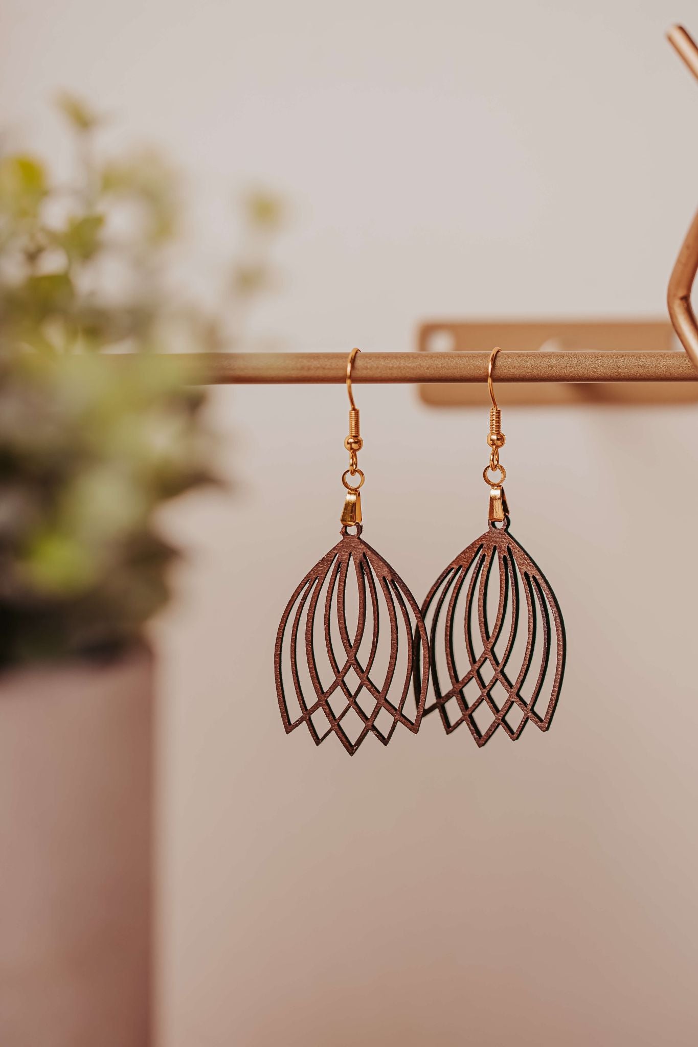 Laser Cut Tulip Earrings by Mirabilia Boutique feature a dark, geometric design and lightweight wooden construction. Hanging from gold rods, these boho statement pieces have hypoallergenic hooks and a softly blurred green foliage background for an elegant touch.