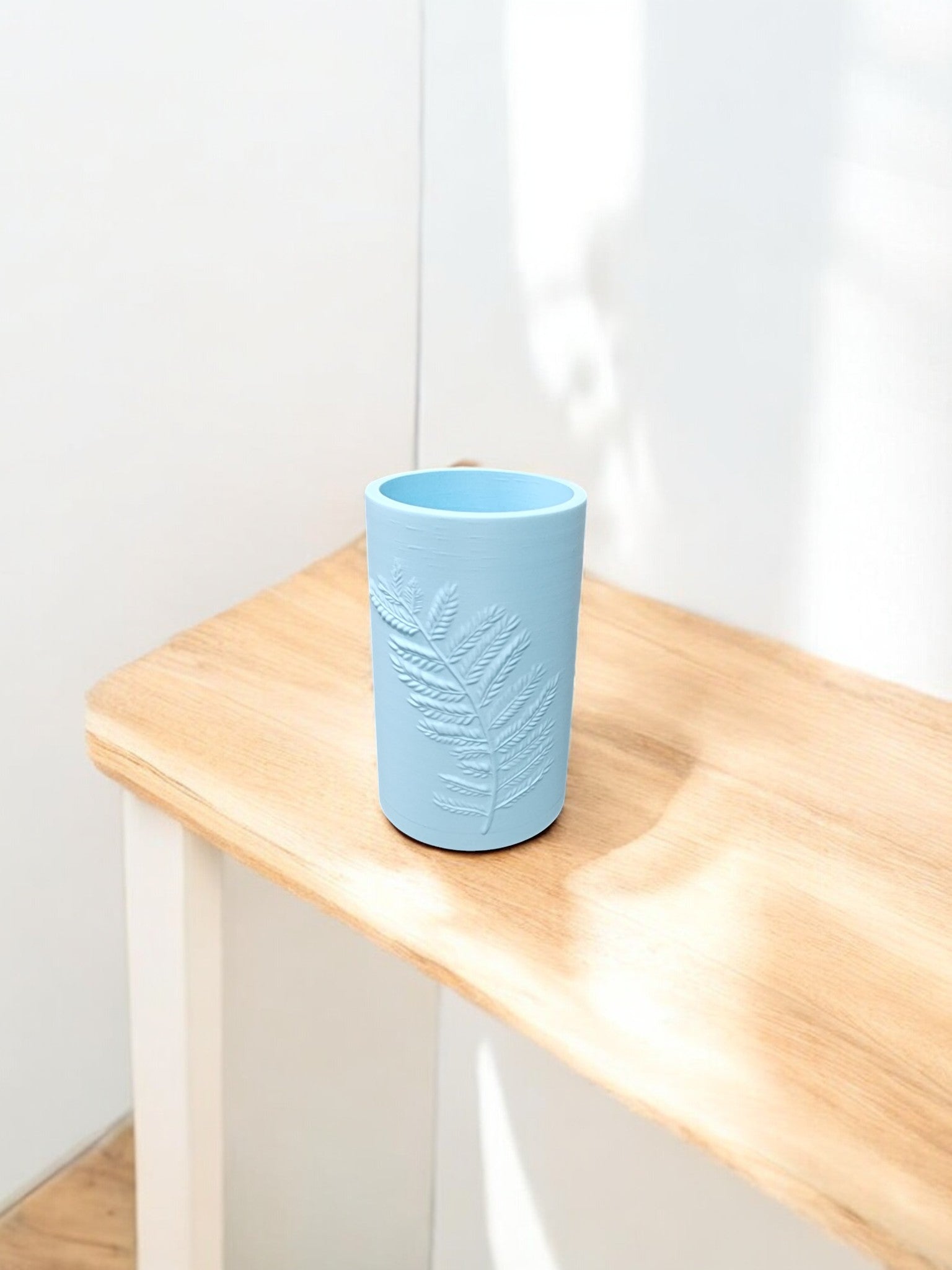 The Mirabilia Boutique Glacial Blue Winter Fern Vase – Woodland Accent features a raised leaf design and sits on a light wooden table in a bright, white-walled room.