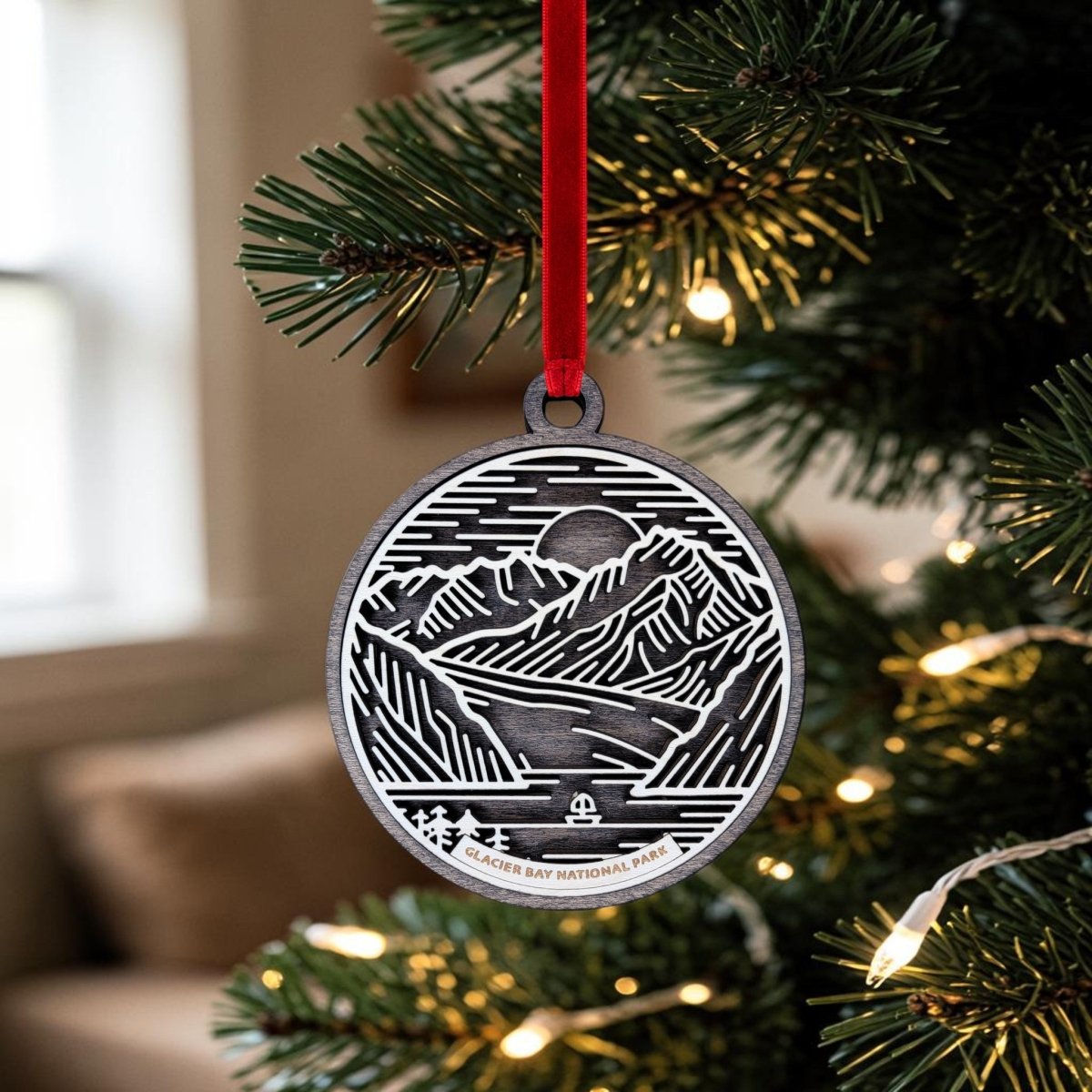 The Glacier Bay National Park Line Art Ornament by Mirabilia Boutique features a silver design engraved with a mountain and glacier scene inspired by the park. Hanging from a red ribbon amidst warm white Christmas lights and a softly blurred indoor background, this ornament is the perfect Alaska keepsake.
