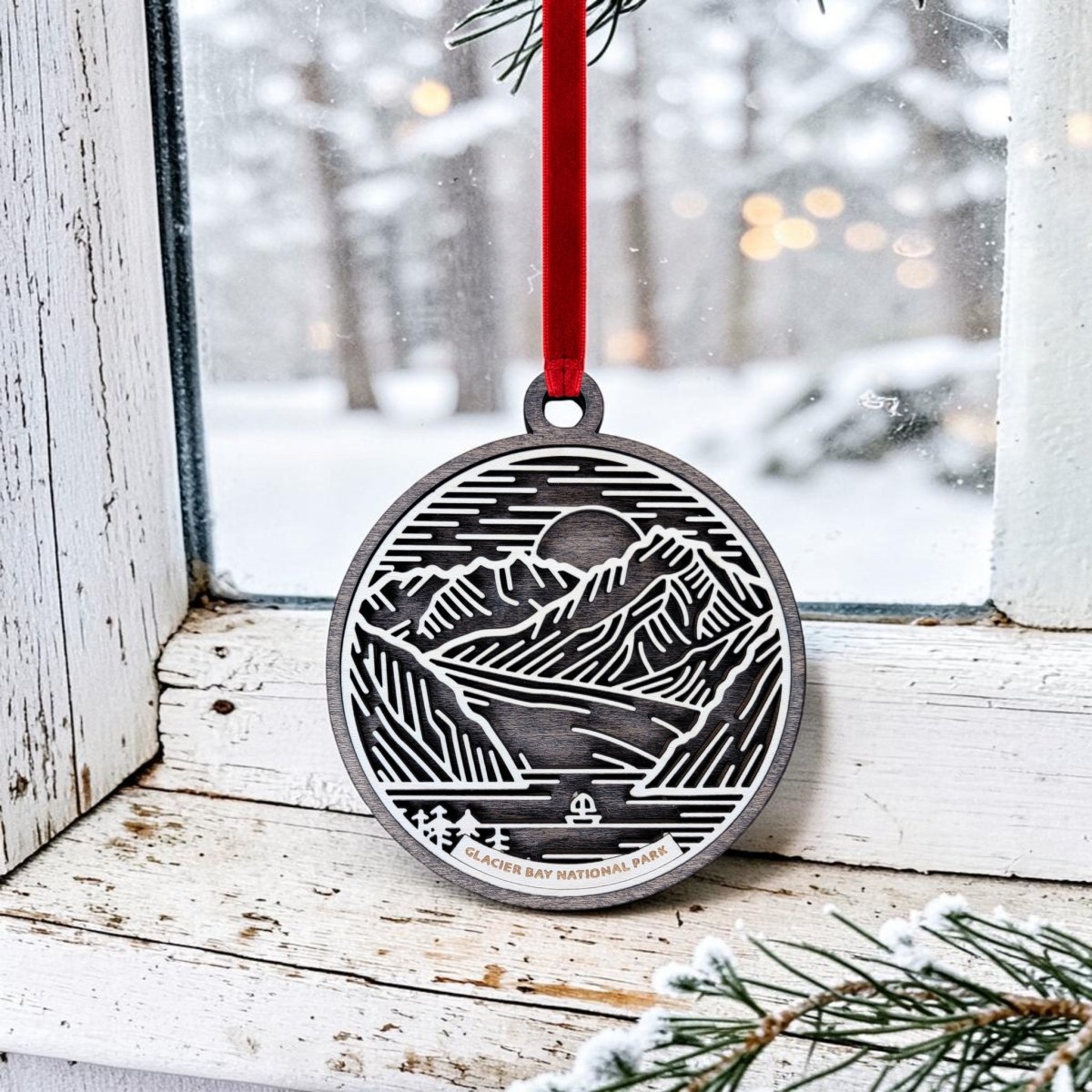 The Glacier Bay National Park Line Art Ornament by Mirabilia Boutique features an oval wooden design with a mountain and glacier motif, labeled "Glacier Bay National Park." Hanging from a red ribbon in a snowy window scene with softly glowing lights and blurred trees outside, this charming piece makes an ideal Alaska keepsake for any holiday décor.