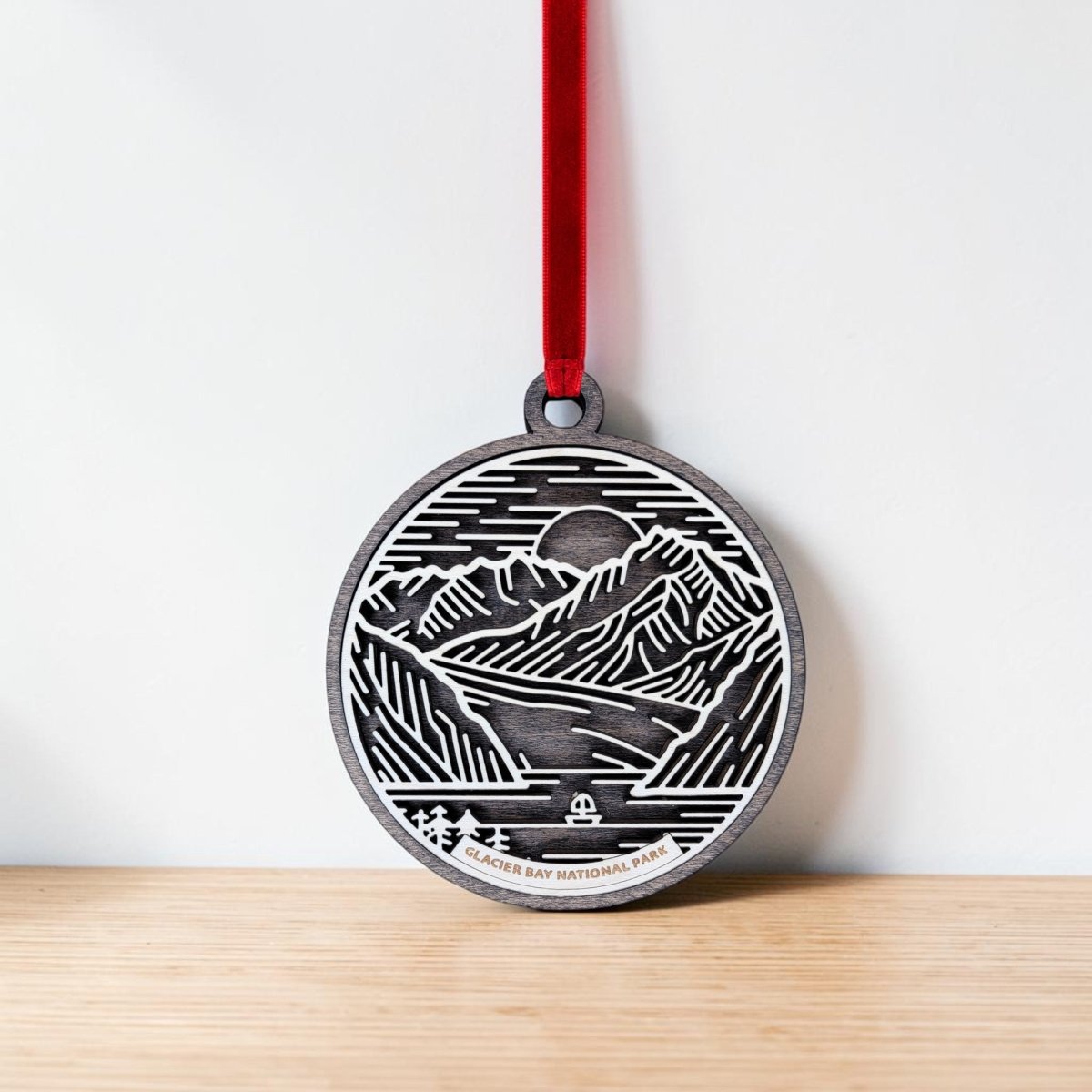 The Glacier Bay National Park Line Art Ornament by Mirabilia Boutique features a round wooden design engraved with a mountain and water scene, finished with "Glacier Bay National Park" lettering. Hanging from a red ribbon against a white wall and wooden surface, it makes an ideal Alaska keepsake for any nature lover.