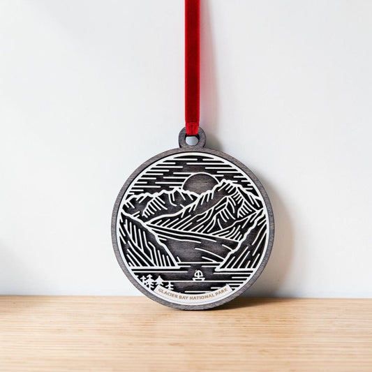 The Glacier Bay National Park Line Art Ornament by Mirabilia Boutique features a round wooden design engraved with a mountain and water scene, finished with "Glacier Bay National Park" lettering. Hanging from a red ribbon against a white wall and wooden surface, it makes an ideal Alaska keepsake for any nature lover.