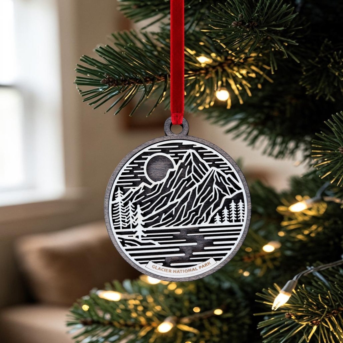 The Glacier National Park Line Art Christmas Ornament by Mirabilia Boutique, featuring a mountain and river design, hangs on a Christmas tree branch adorned with lights, bringing a charming wooden keepsake touch to your holiday decor.