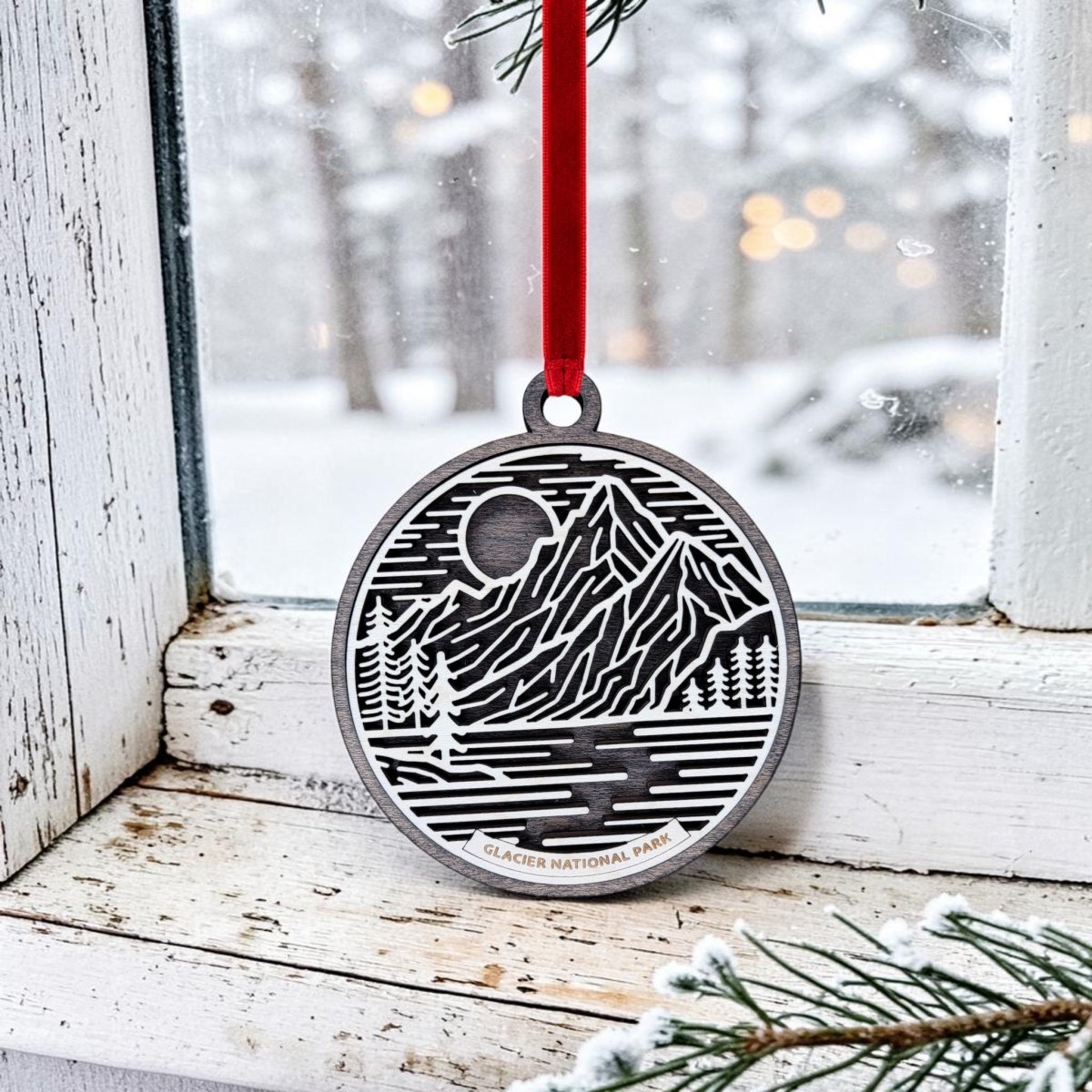The Mirabilia Boutique Glacier National Park Line Art Christmas Ornament is an oval wooden keepsake featuring a black and white line art design of mountains, river, and trees with the label "Glacier National Park." It hangs by a red ribbon on a snowy window sill, with a pine branch visible in the upper right corner.