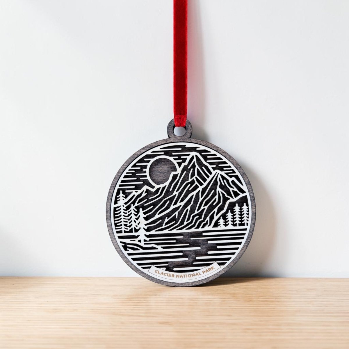 The Glacier National Park Line Art Christmas Ornament by Mirabilia Boutique features delicate line art of mountains, trees, a moon, and a river, all beautifully rendered on a round wooden ornament. "Glacier National Park" is inscribed at the bottom, and the ornament hangs from a red ribbon. This charming keepsake is shown resting against a light wall on a wooden surface.