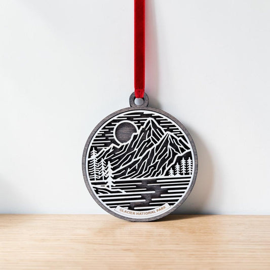 The Glacier National Park Line Art Christmas Ornament by Mirabilia Boutique features delicate line art of mountains, trees, a moon, and a river, all beautifully rendered on a round wooden ornament. "Glacier National Park" is inscribed at the bottom, and the ornament hangs from a red ribbon. This charming keepsake is shown resting against a light wall on a wooden surface.