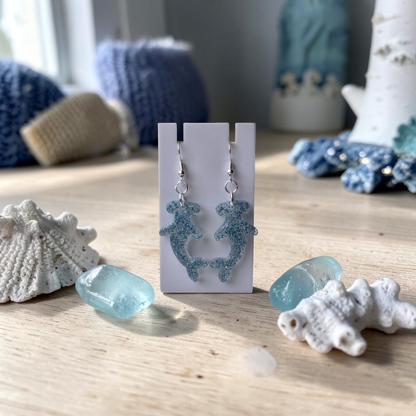 The Blue Glitter Hammerhead Shark Earrings by Mirabilia Boutique are displayed on a white stand, surrounded by seashells and light blue sea glass—perfect ocean-inspired jewelry for any look.