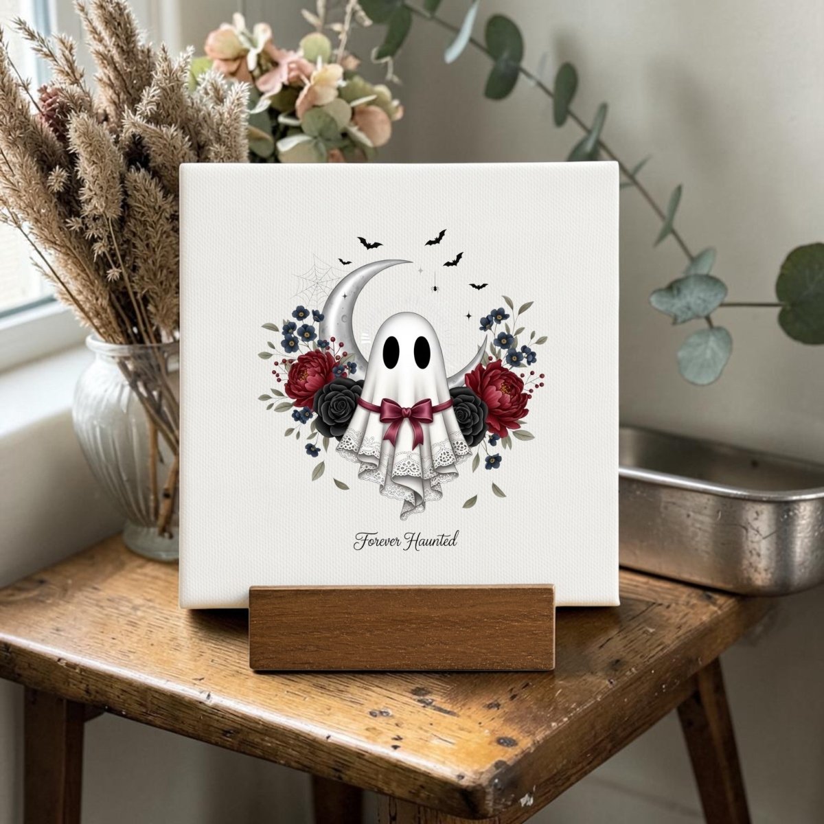 The Forever Haunted Mini Canvas Sign by Mirabilia Boutique features a charming ghost in a veil amid roses, leaves, and bats, with the phrase “Forever Haunted.” Displayed on a wooden stand atop a rustic table near a window adorned with dried flowers, this piece adds the perfect gothic cottagecore accent to your Halloween home.