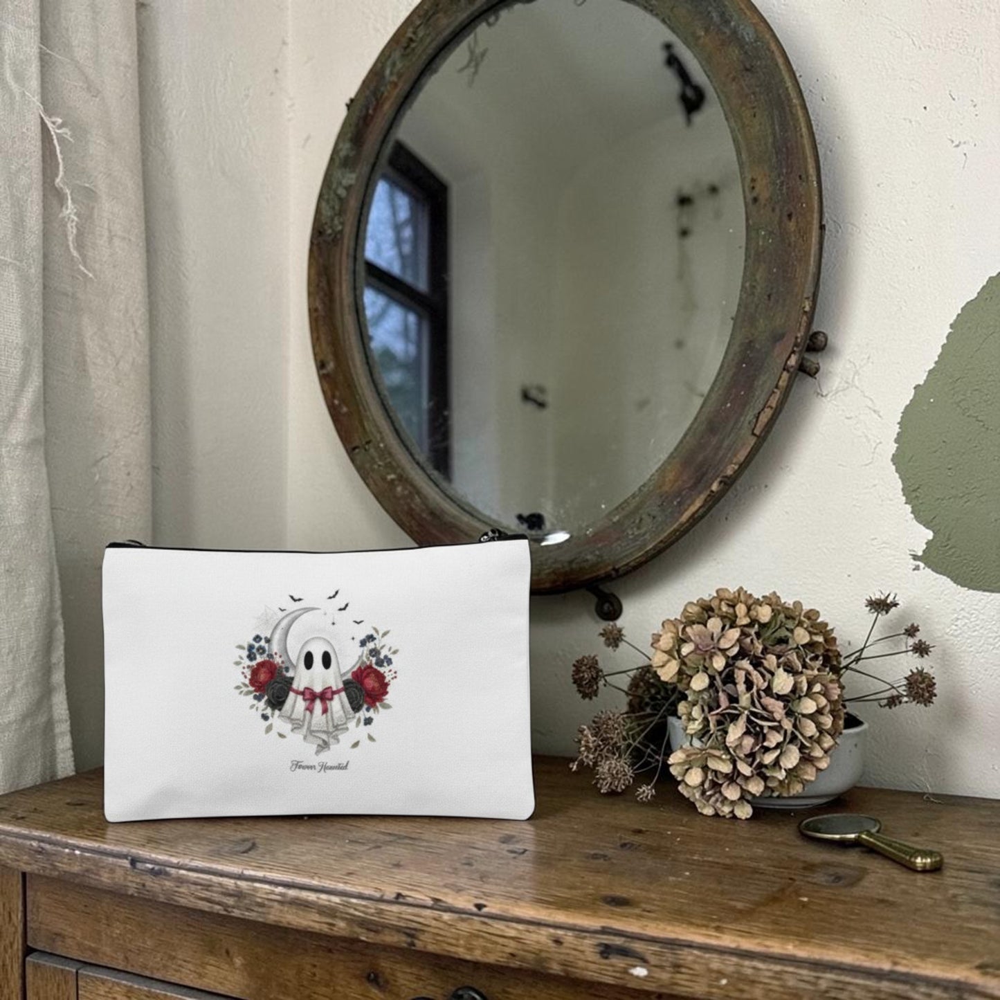 The Forever Haunted Makeup Bag | Gothic Ghost Pouch by Mirabilia Boutique—a small white gothic cosmetic pouch adorned with a skull and floral design—rests on a wooden dresser. Beside it lie dried flowers, an old-fashioned key, and a round antique mirror reflecting a window.