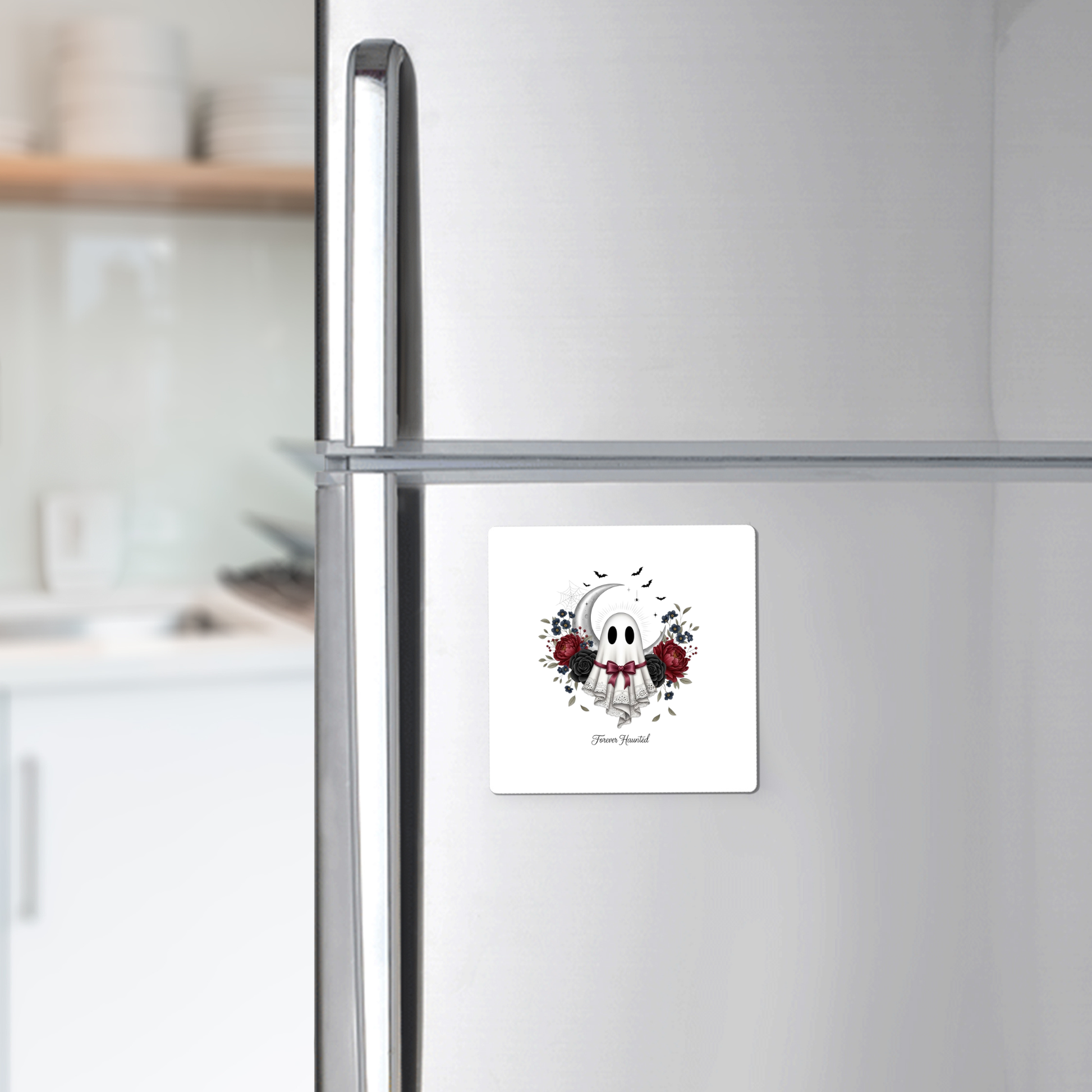 A silver refrigerator displays the Mirabilia Boutique "Forever Haunted Ghost Magnet | Gothic Halloween," a square magnet featuring a cute ghost, red and black roses, and text along the lower edge. Set against a blurred white kitchen with shelves and dishes, this gothic Halloween décor piece stands out beautifully.