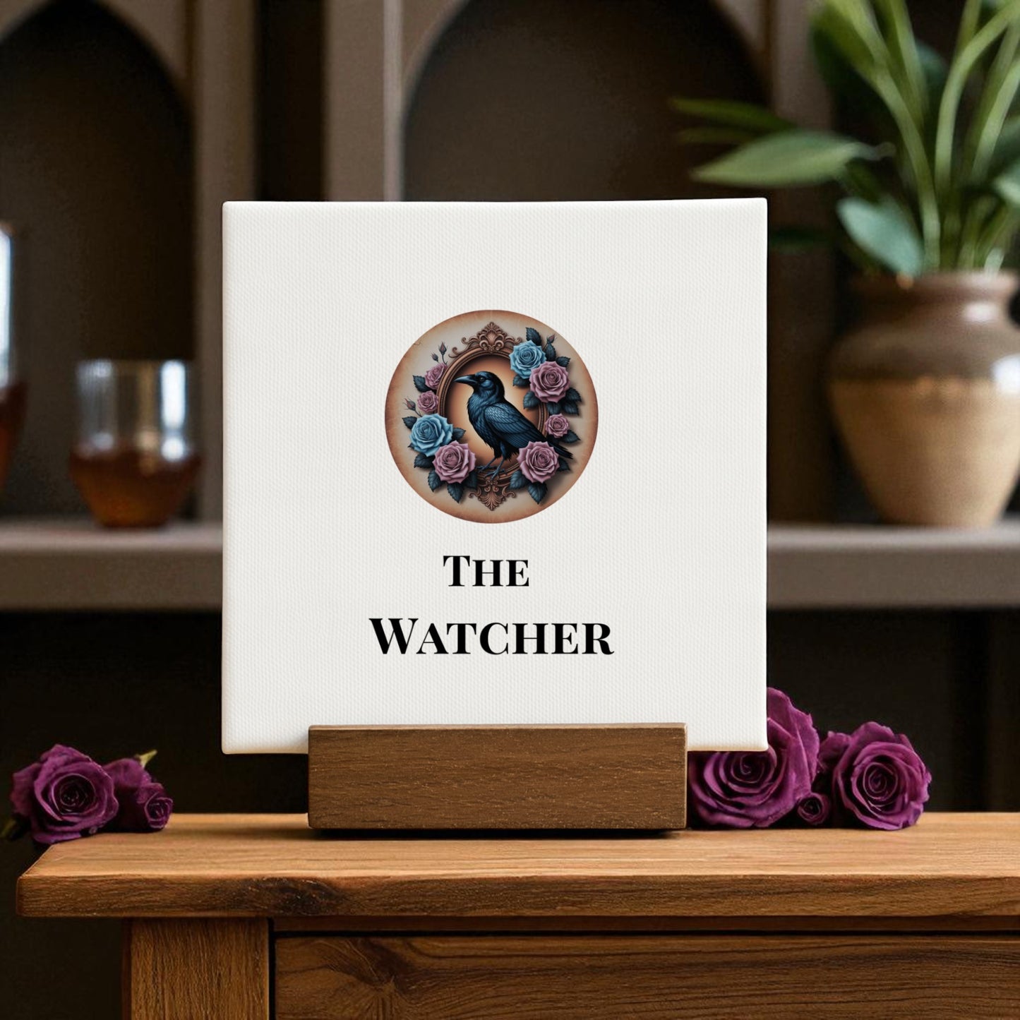 The Mini Gothic Sign | The Watcher by Mirabilia Boutique displays a black raven amid purple roses on a white square, perched on a wooden stand. Set against gothic decor with greenery, it brings raven symbolism to your space.