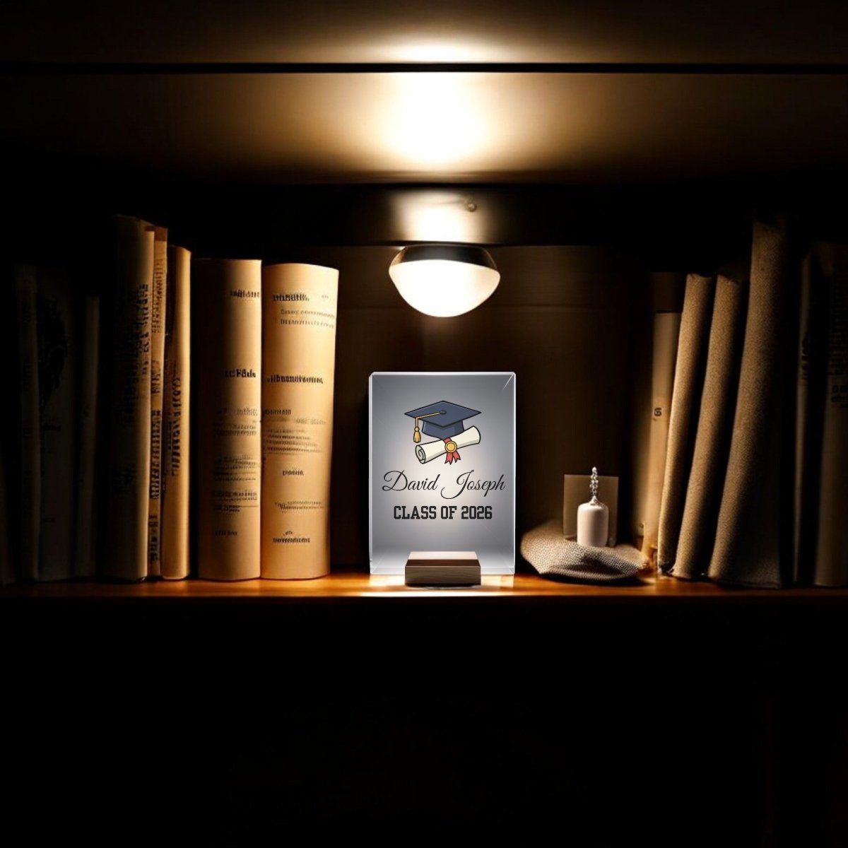 A bookshelf glows with the Mirabilia Boutique Classic Graduation Cap Personalized Night Light and features a custom graduation plaque for “David Joseph, Class of 2026,” books, a bottle, and folded cloth nearby.