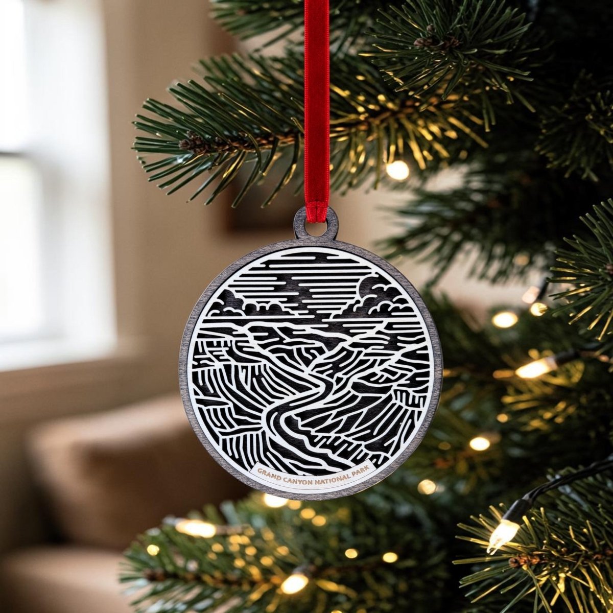 The Grand Canyon National Park Line Art Ornament by Mirabilia Boutique, a charming wooden keepsake featuring a black and white landscape design, hangs from a red ribbon on a Christmas tree adorned with lights.