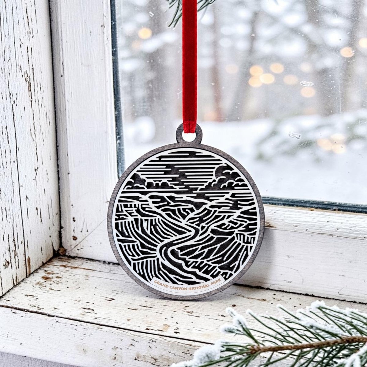 A Grand Canyon National Park Line Art Ornament from Mirabilia Boutique, featuring a black and white landscape design, hangs by a red ribbon on a snow-dusted window, making for a charming wooden keepsake against the wintry backdrop of snowy trees outside.