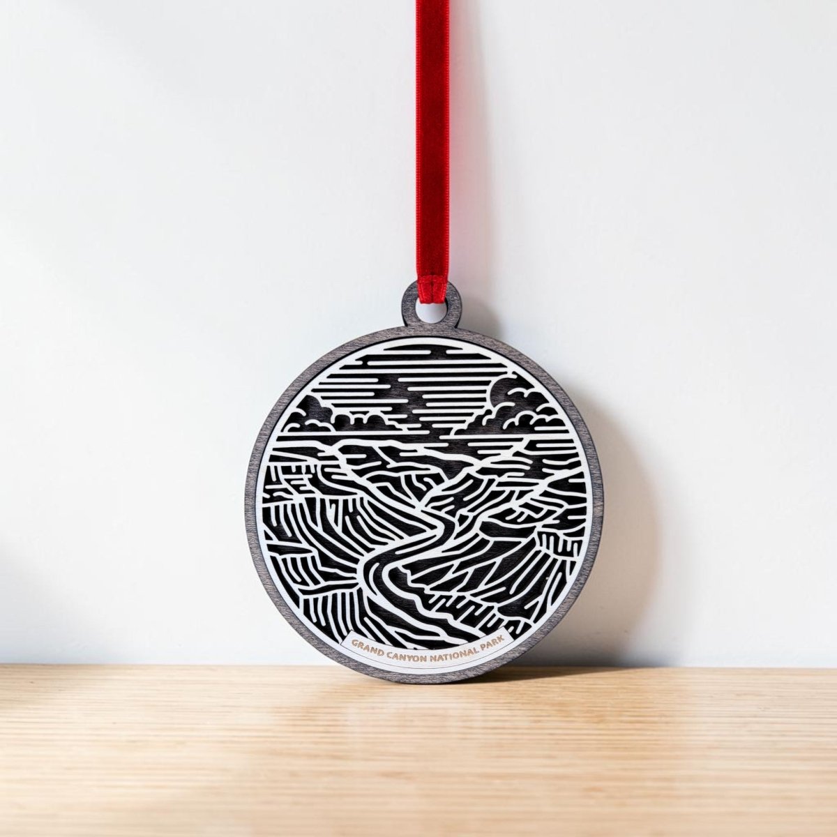The Grand Canyon National Park Line Art Ornament by Mirabilia Boutique, a round wooden keepsake featuring a black and white Grand Canyon design, hangs from a red ribbon against a white wall above a light wood surface.