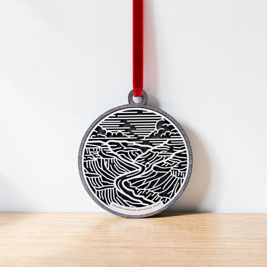 The Grand Canyon National Park Line Art Ornament by Mirabilia Boutique, a round wooden keepsake featuring a black and white Grand Canyon design, hangs from a red ribbon against a white wall above a light wood surface.