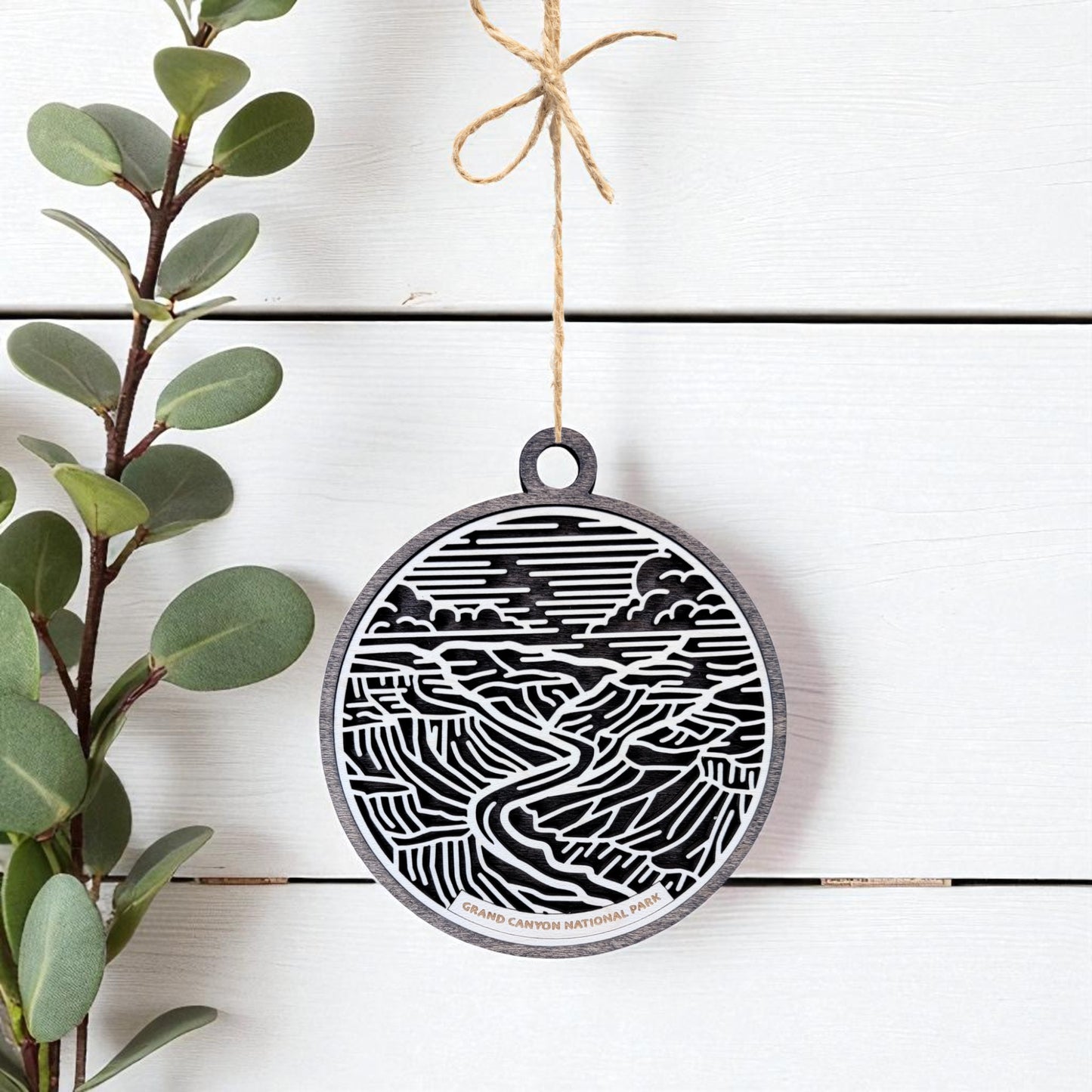 The Grand Canyon National Park Ornament by Mirabilia Boutique, a round wooden travel gift engraved with a canyon landscape, hangs from twine on a white background with green leaves—a unique national park keepsake.