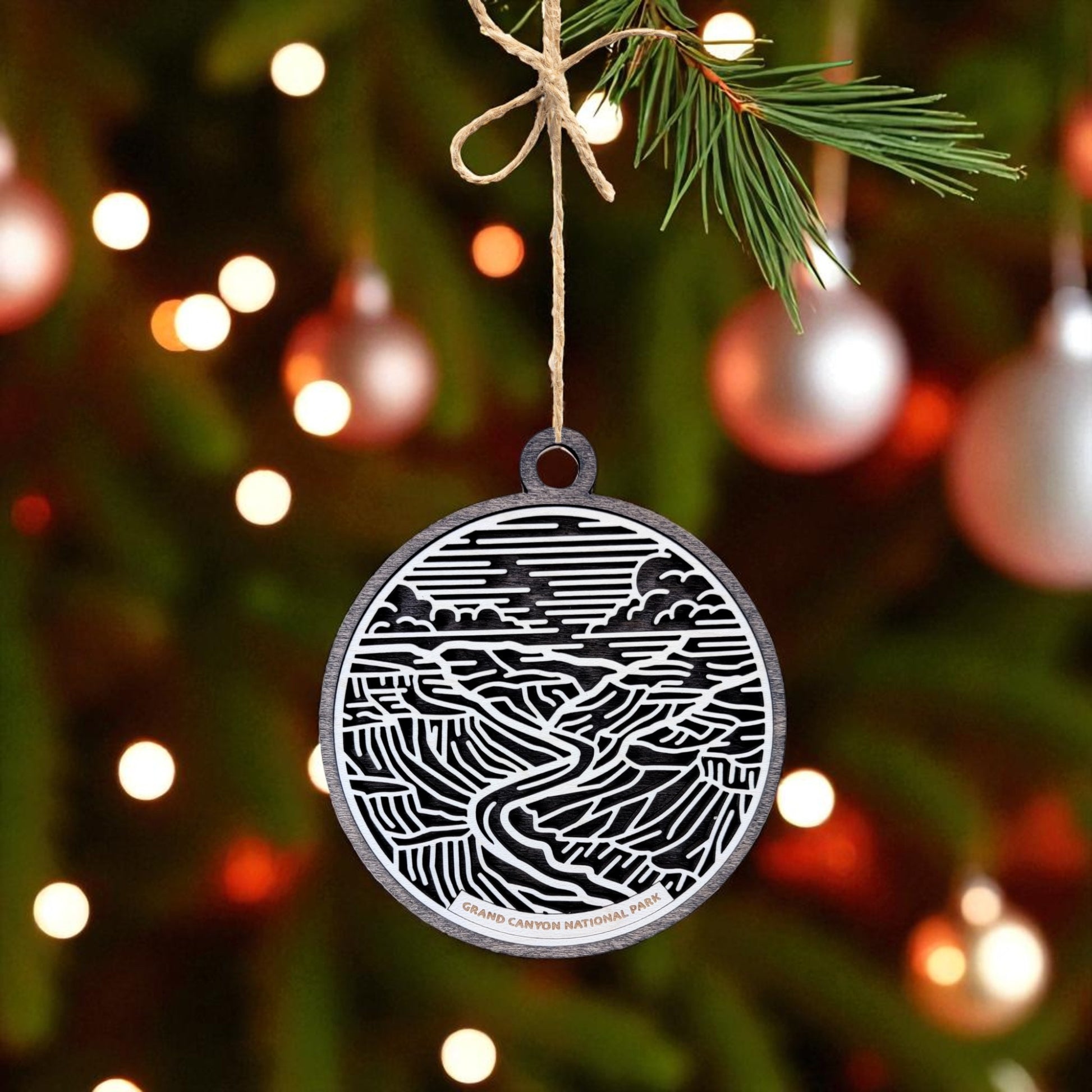The Grand Canyon National Park Ornament by Mirabilia Boutique, a round wooden travel gift featuring a black and white line design, hangs from a pine branch with festive lights—a perfect holiday gift for national park lovers.