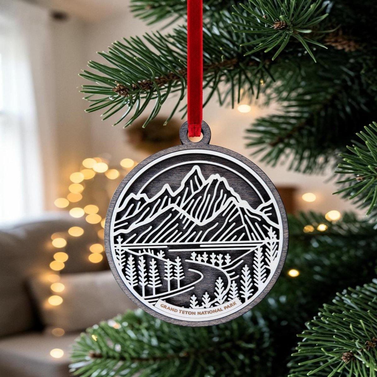 The Grand Teton National Park Line Art Ornament by Mirabilia Boutique, featuring a mountain and road design in metal, hangs from a red ribbon on a Christmas tree. Softly blurred warm lights and a sofa can be seen in the background.