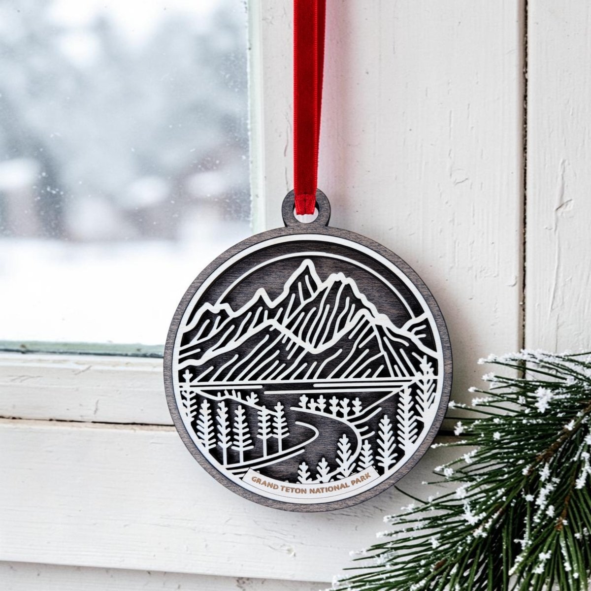A Grand Teton National Park Line Art Ornament by Mirabilia Boutique—oval-shaped and crafted from wood with a cutout design featuring mountains, trees, and a winding road—hangs from a red ribbon near a snowy window, while pine needles are visible in the bottom right corner.