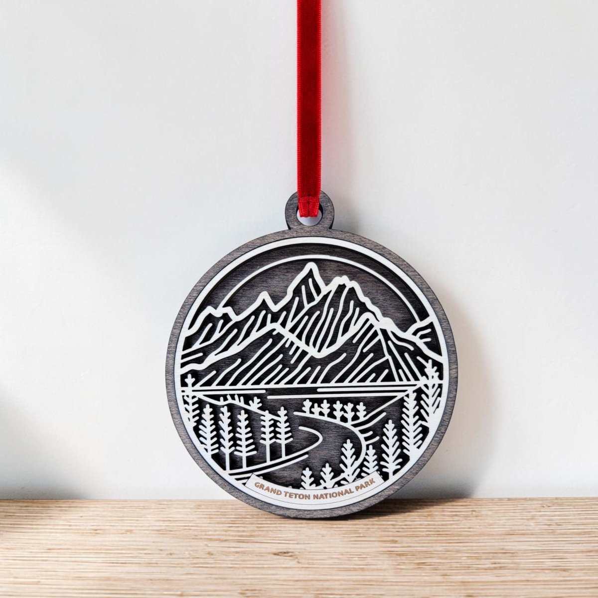 The Mirabilia Boutique Grand Teton National Park Line Art Ornament is a round wooden keepsake featuring a cutout, line art design of mountains, trees, and a road. Hanging by a red ribbon, it displays the text “Grand Teton National Park” at the bottom. In the image, the ornament rests on a wood surface against a white wall.