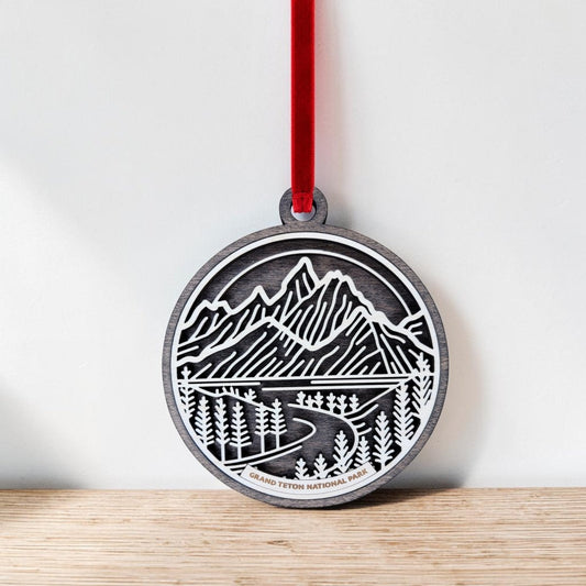 The Mirabilia Boutique Grand Teton National Park Line Art Ornament is a round wooden keepsake featuring a cutout, line art design of mountains, trees, and a road. Hanging by a red ribbon, it displays the text “Grand Teton National Park” at the bottom. In the image, the ornament rests on a wood surface against a white wall.