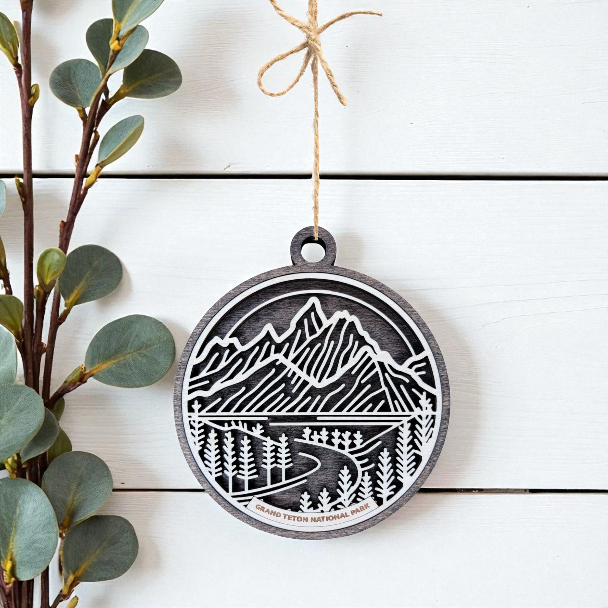 The Grand Teton National Park Ornament by Mirabilia Boutique features a detailed wooden mountain, tree, and river cutout, labeled "Grand Teton National Park," hanging by twine—ideal for nature-inspired holiday décor.